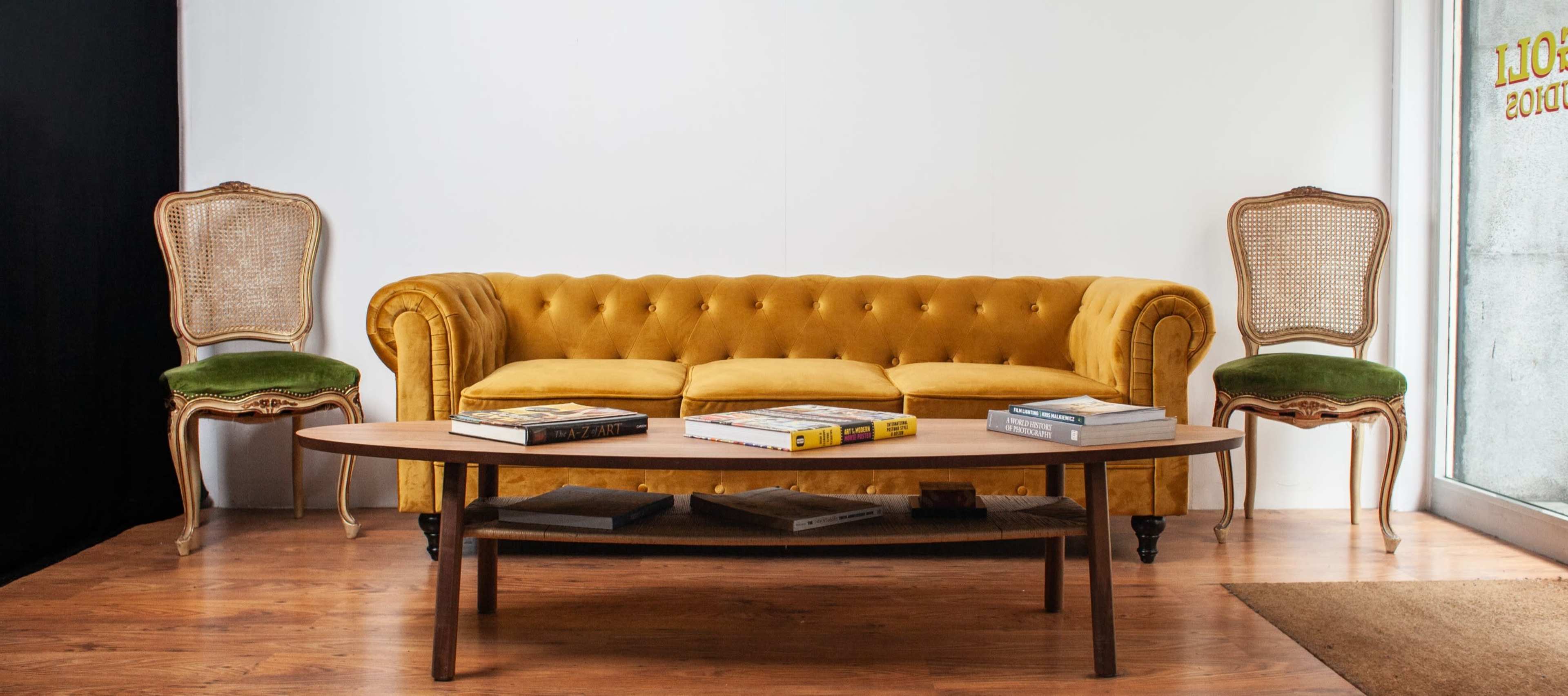 A yellow tufted sofa is positioned in the center of a room, flanked by two ornate chairs and a coffee table displaying books.