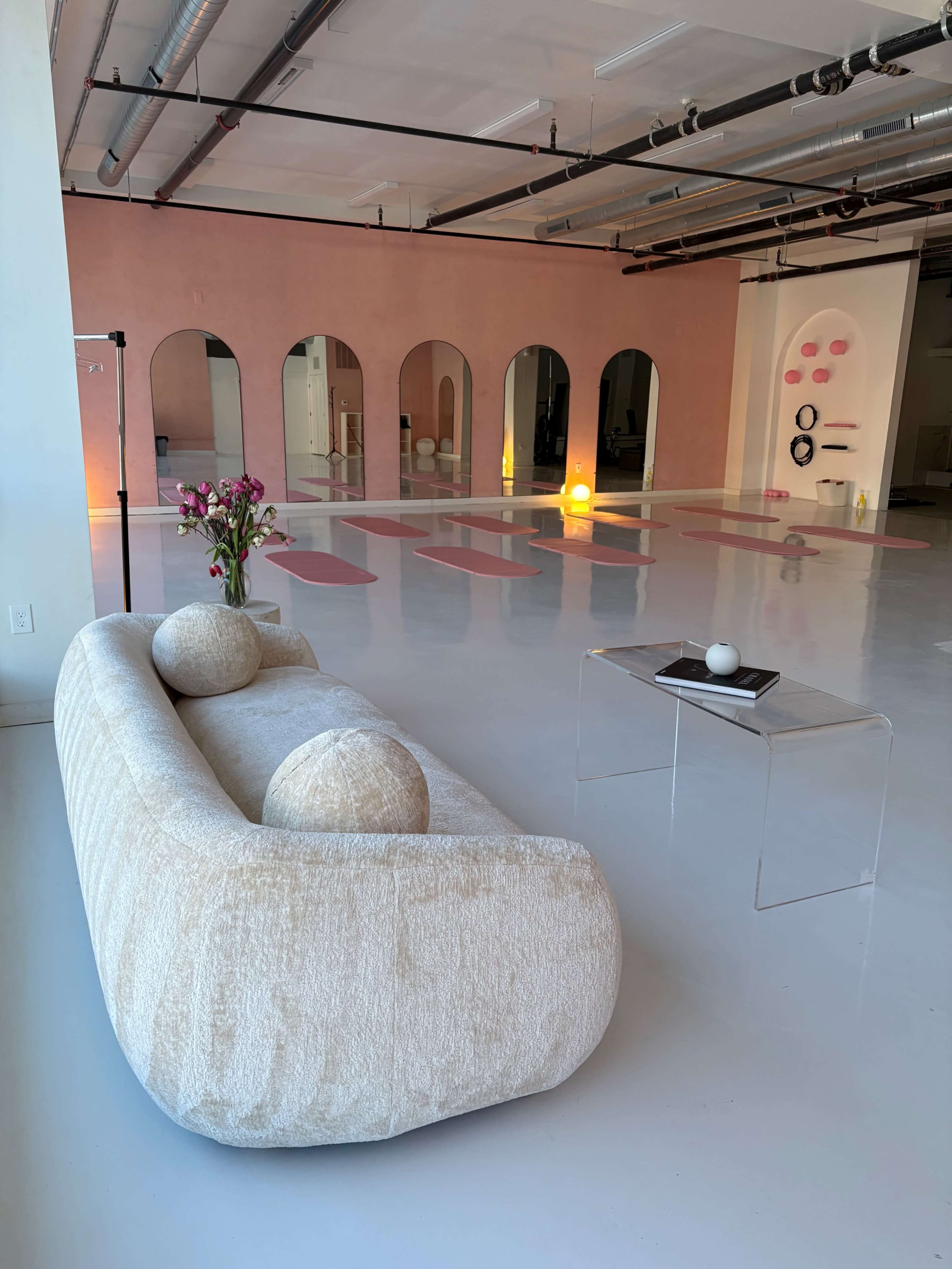 The image shows a spacious room with a light-colored floor, featuring a modern beige sofa with round cushions, a clear side table with a decorative item, and a series of yoga mats arranged in the background.
