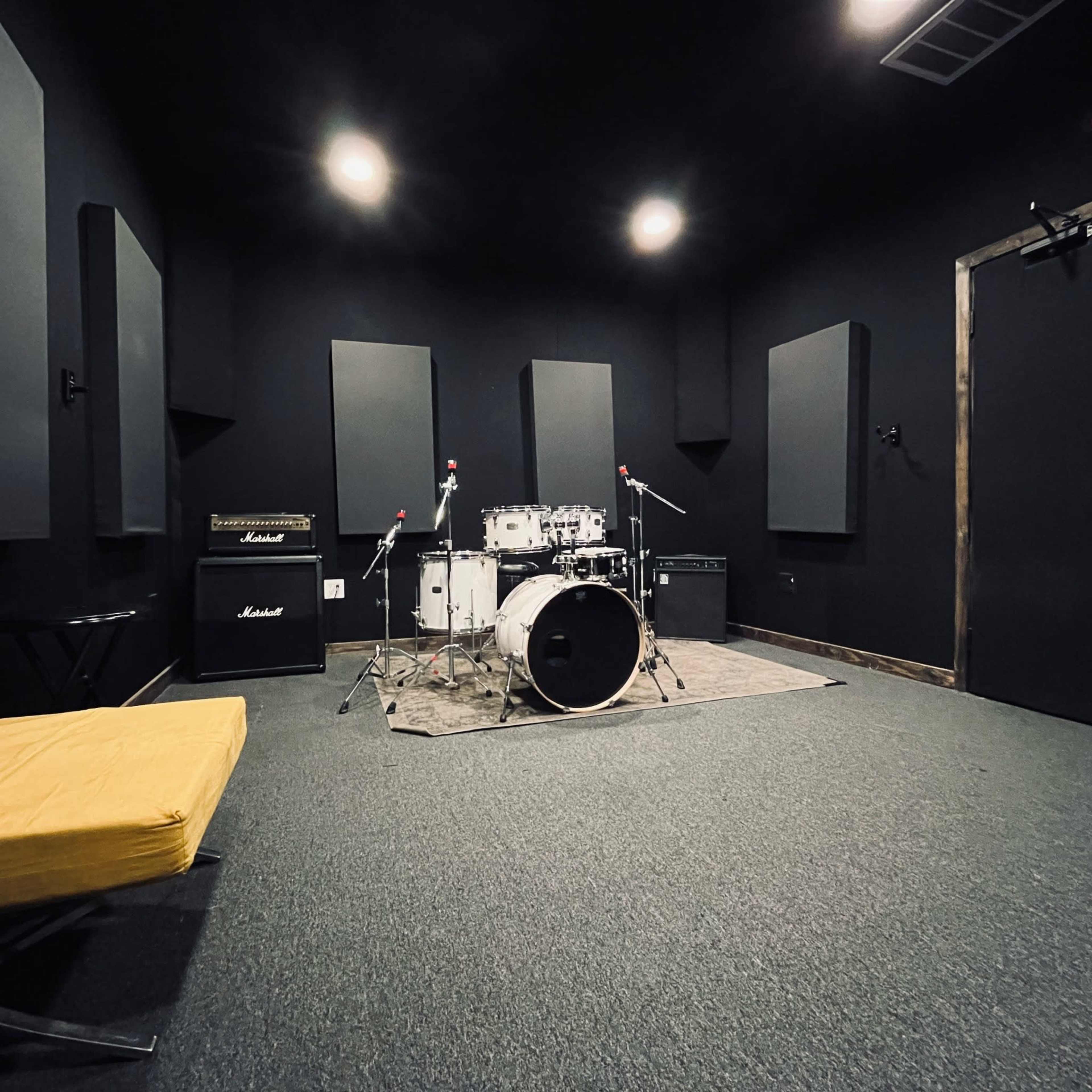 The image shows a music rehearsal room featuring a drum set, amplifiers, and soundproofing panels on the walls.