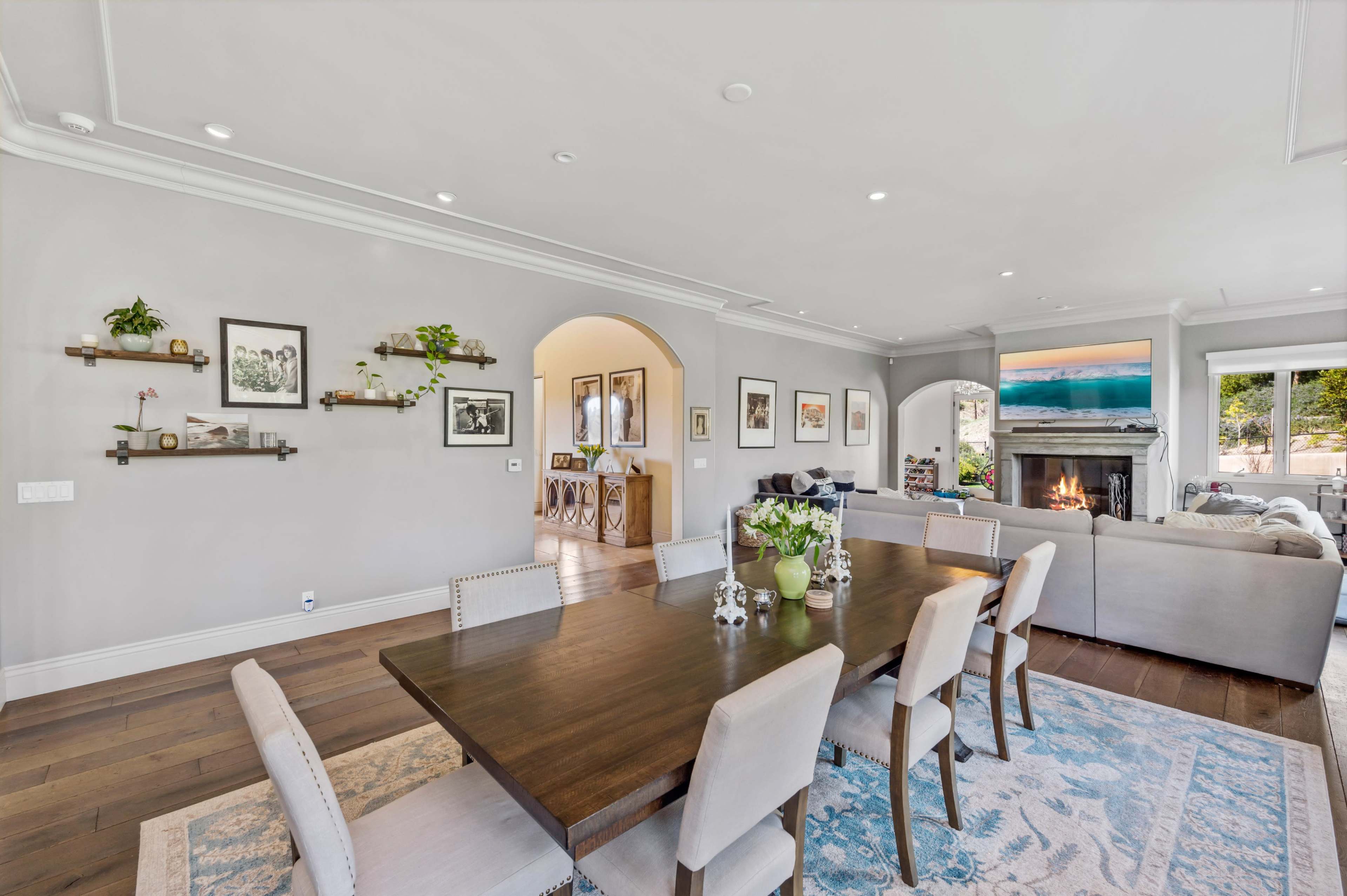 A dining area with a wooden table surrounded by six chairs is adjacent to a cozy living room featuring a fireplace and large windows.