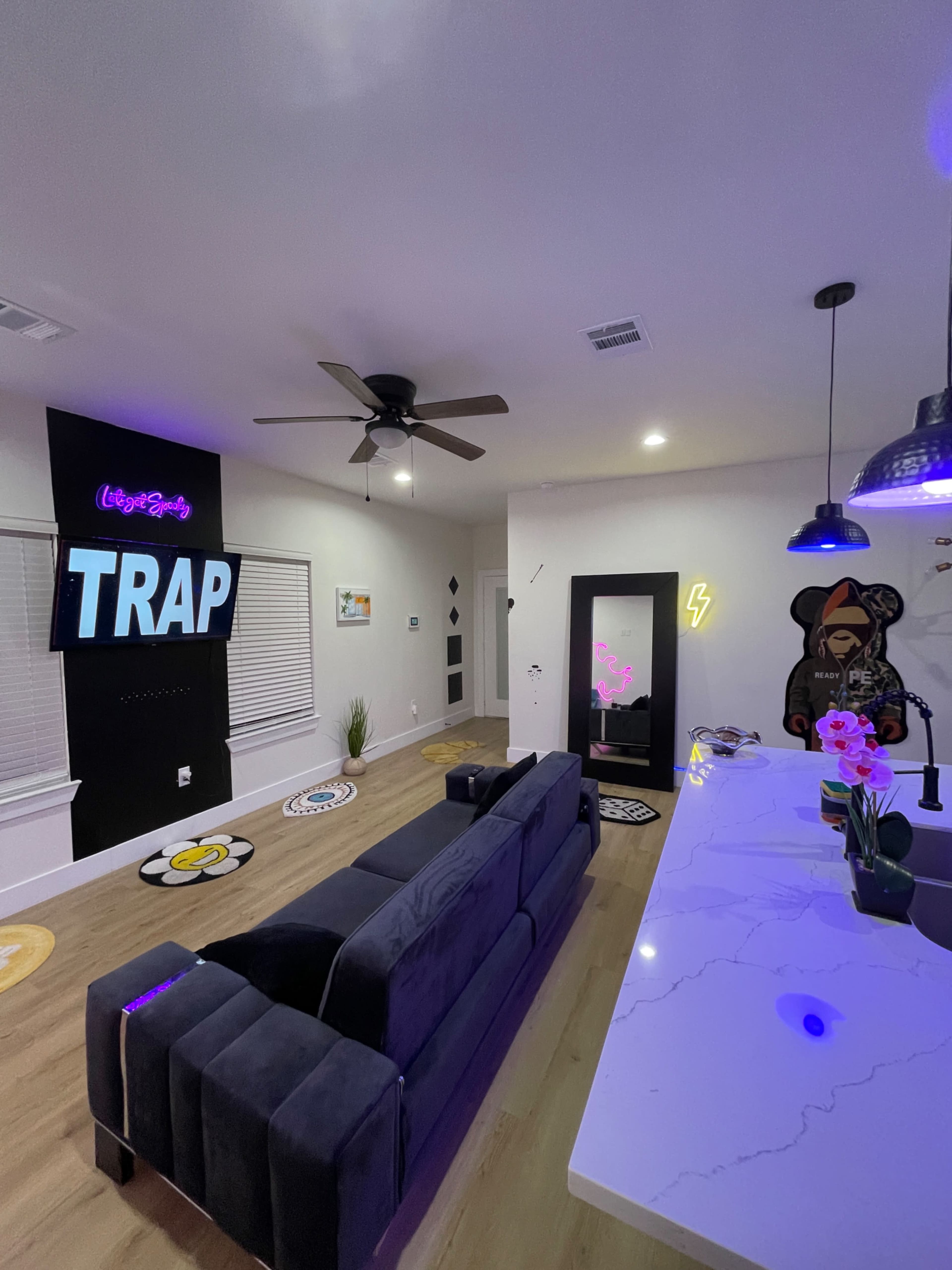 The image shows a modern living room with a gray couch, a wall featuring neon text that reads "TRAP," and a bar area with a sleek countertop under pendant lighting.