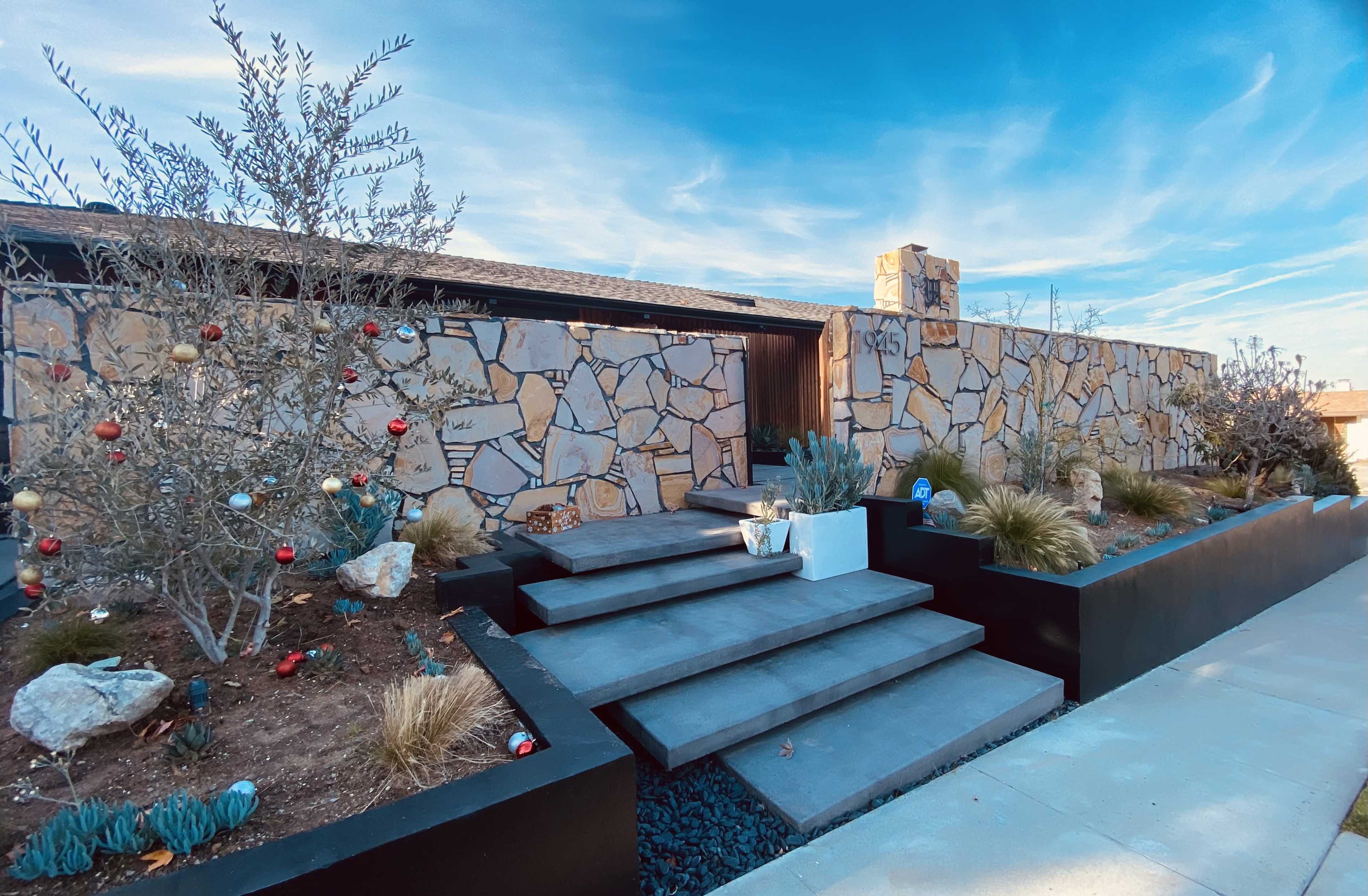 The image shows a modern home entrance with a stone wall, layered steps, and landscaped shrubs and plants.