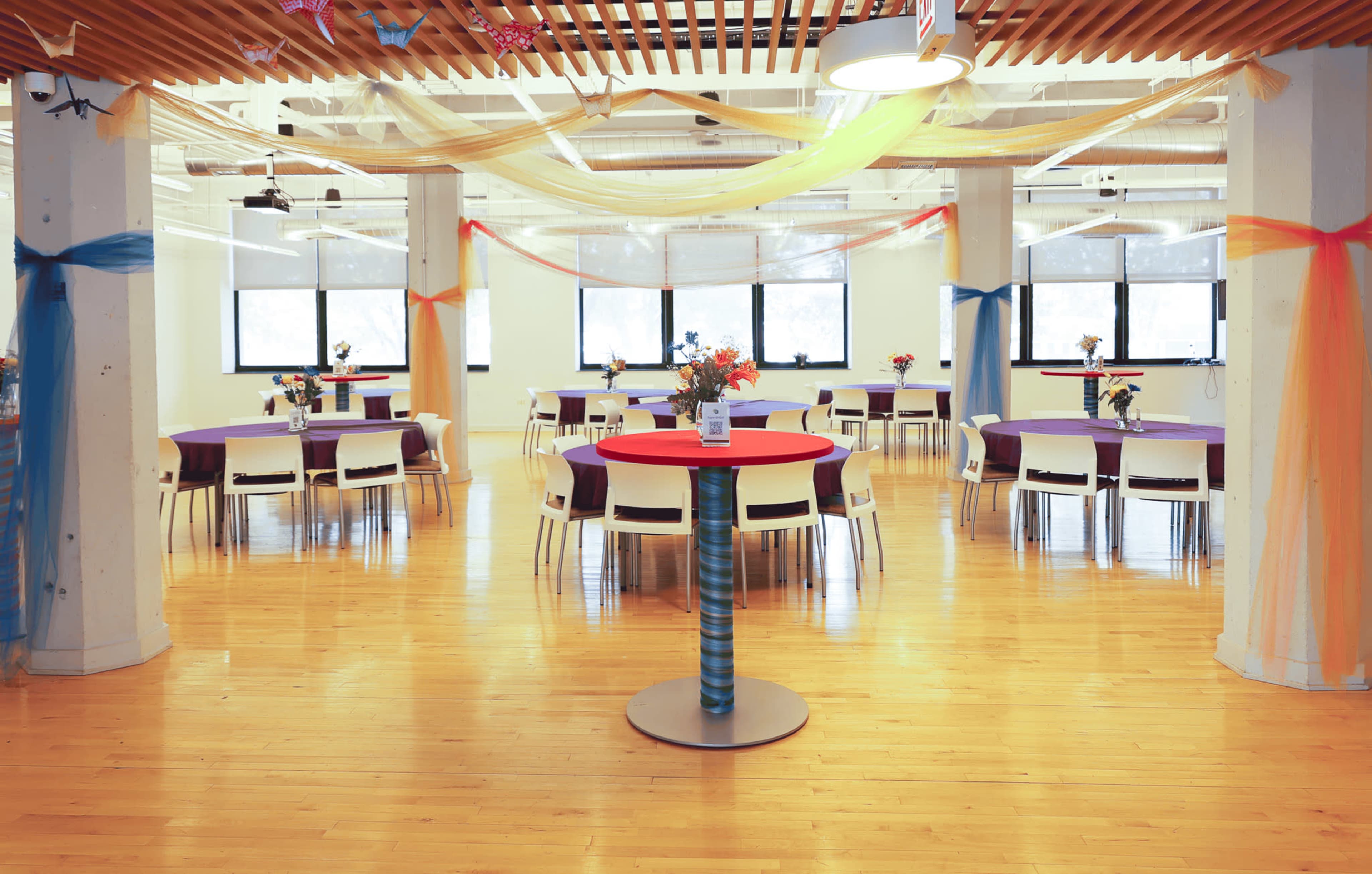 The image shows a spacious event room with decorated tables, featuring flowers and colorful tablecloths, under a ceiling adorned with draped fabric.