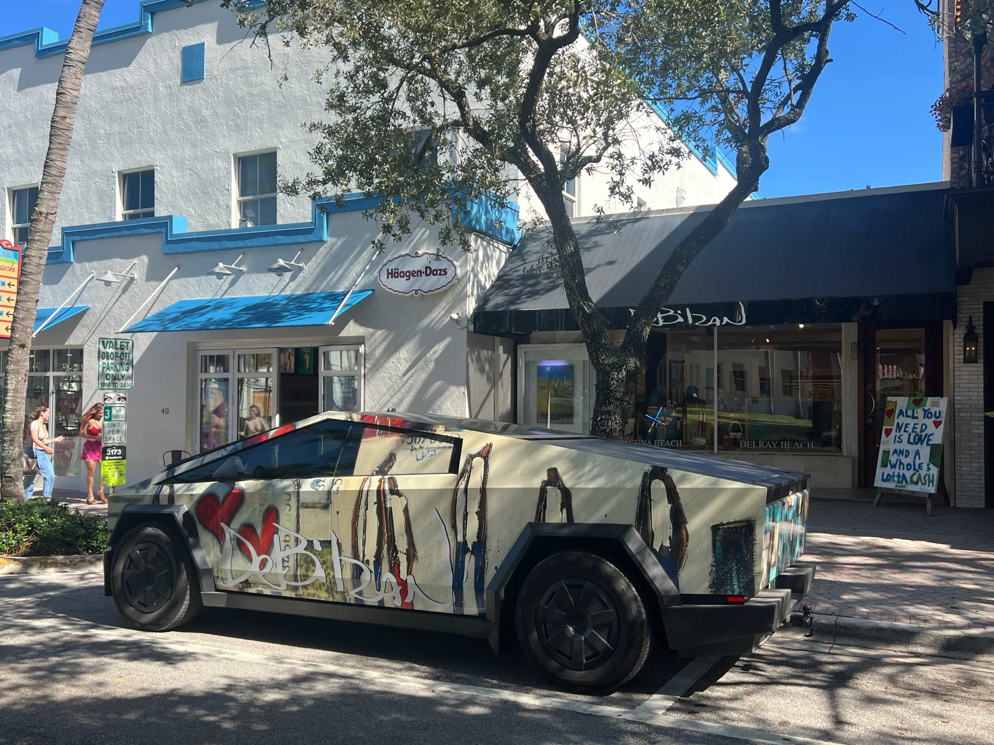 A graffiti-covered Tesla Cybertruck is parked along a tree-lined street beside a store front with large windows.