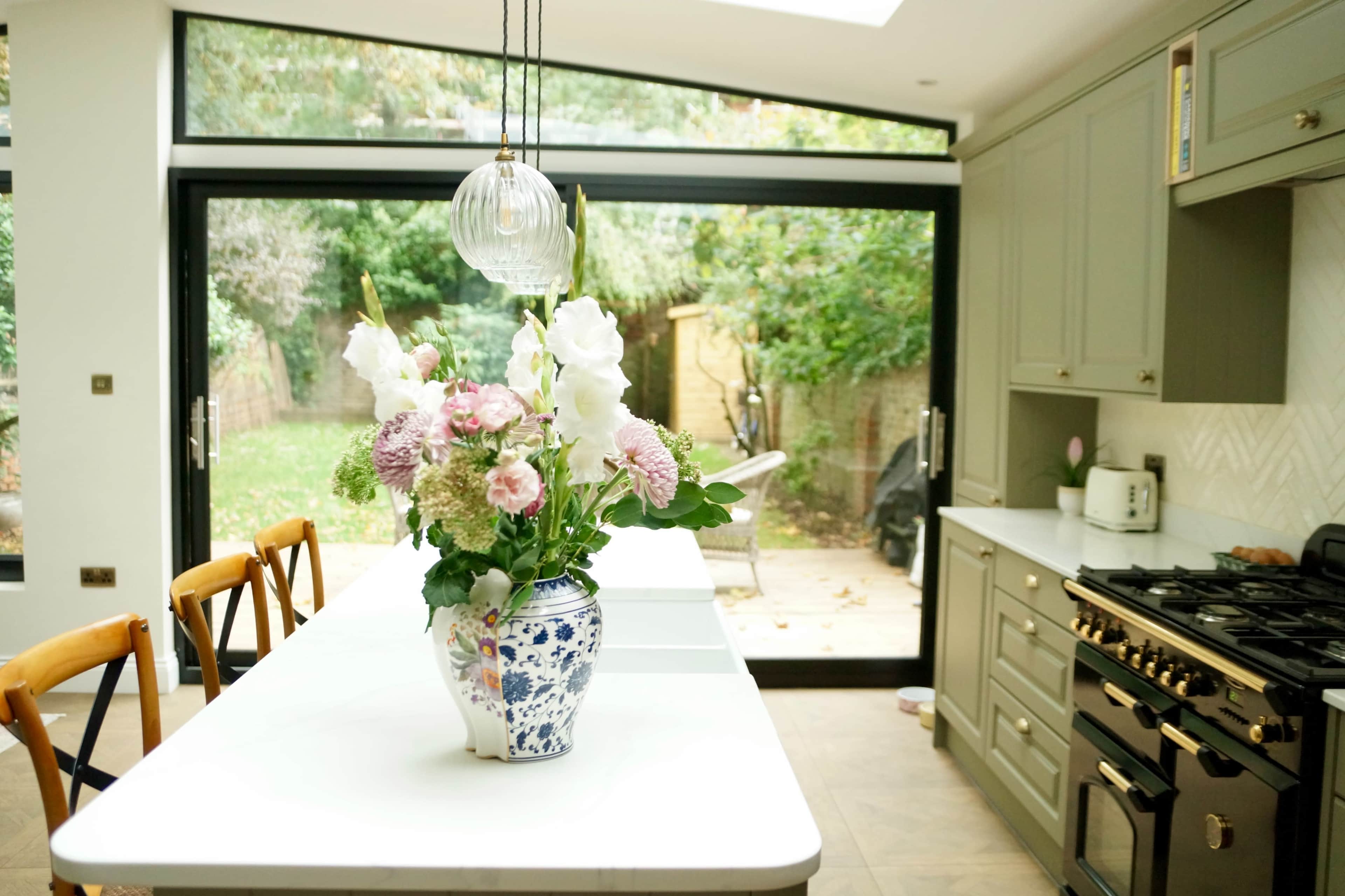 A modern kitchen features a large island with a floral arrangement in a vase, and large glass doors that open to a garden.