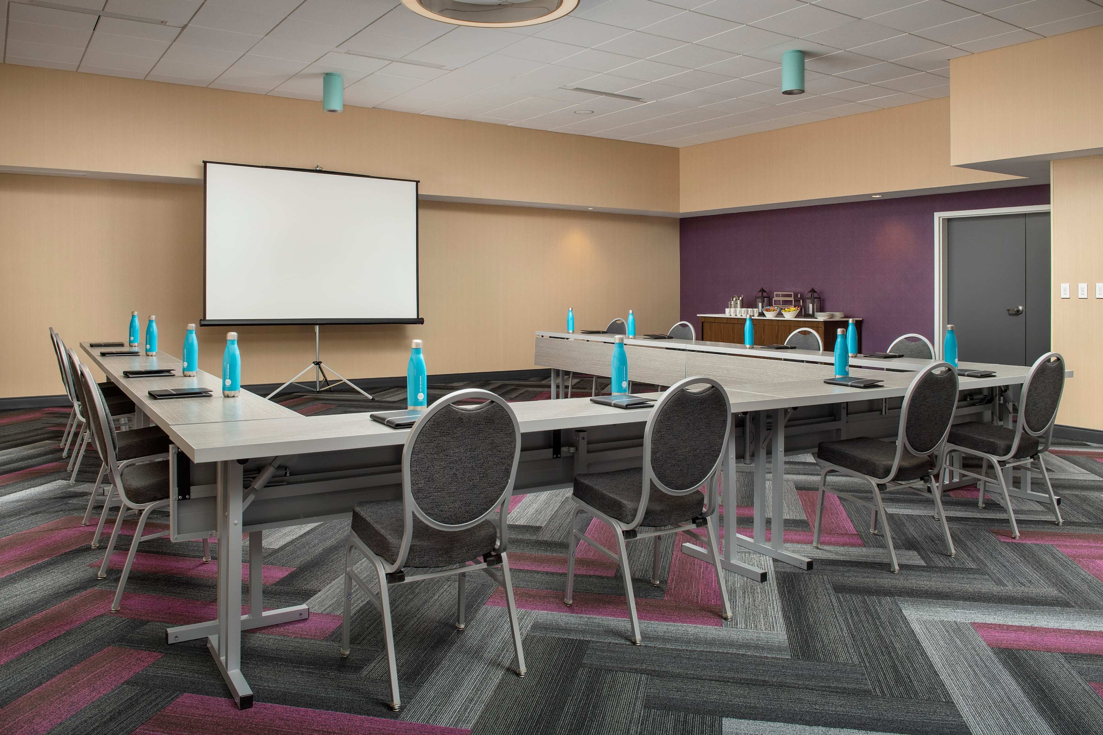 A modern conference room features a long table with chairs, a projection screen, and water bottles at each place setting.