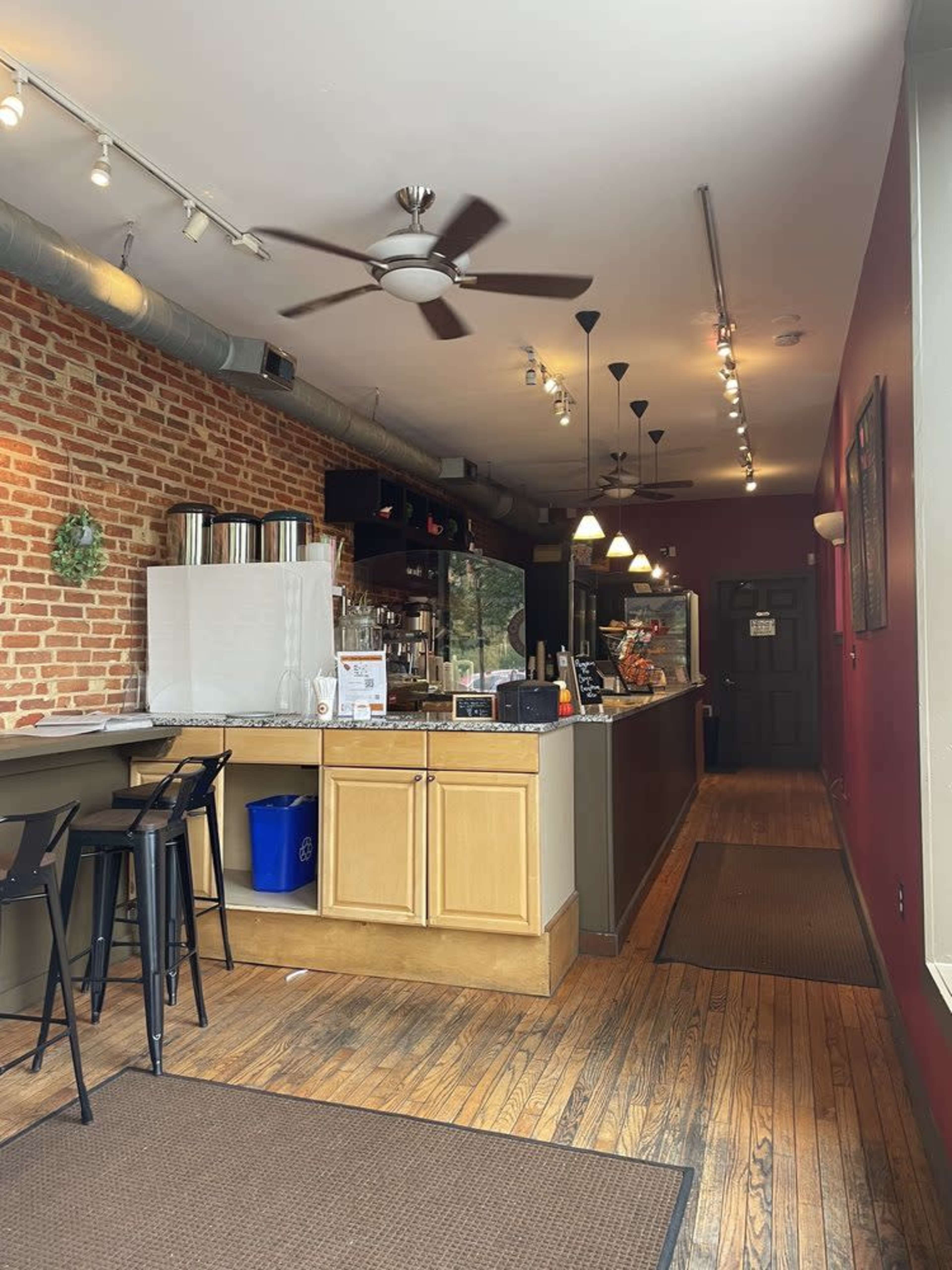 The image shows the interior of a café featuring a brick wall, a counter with seating, and a fan overhead.