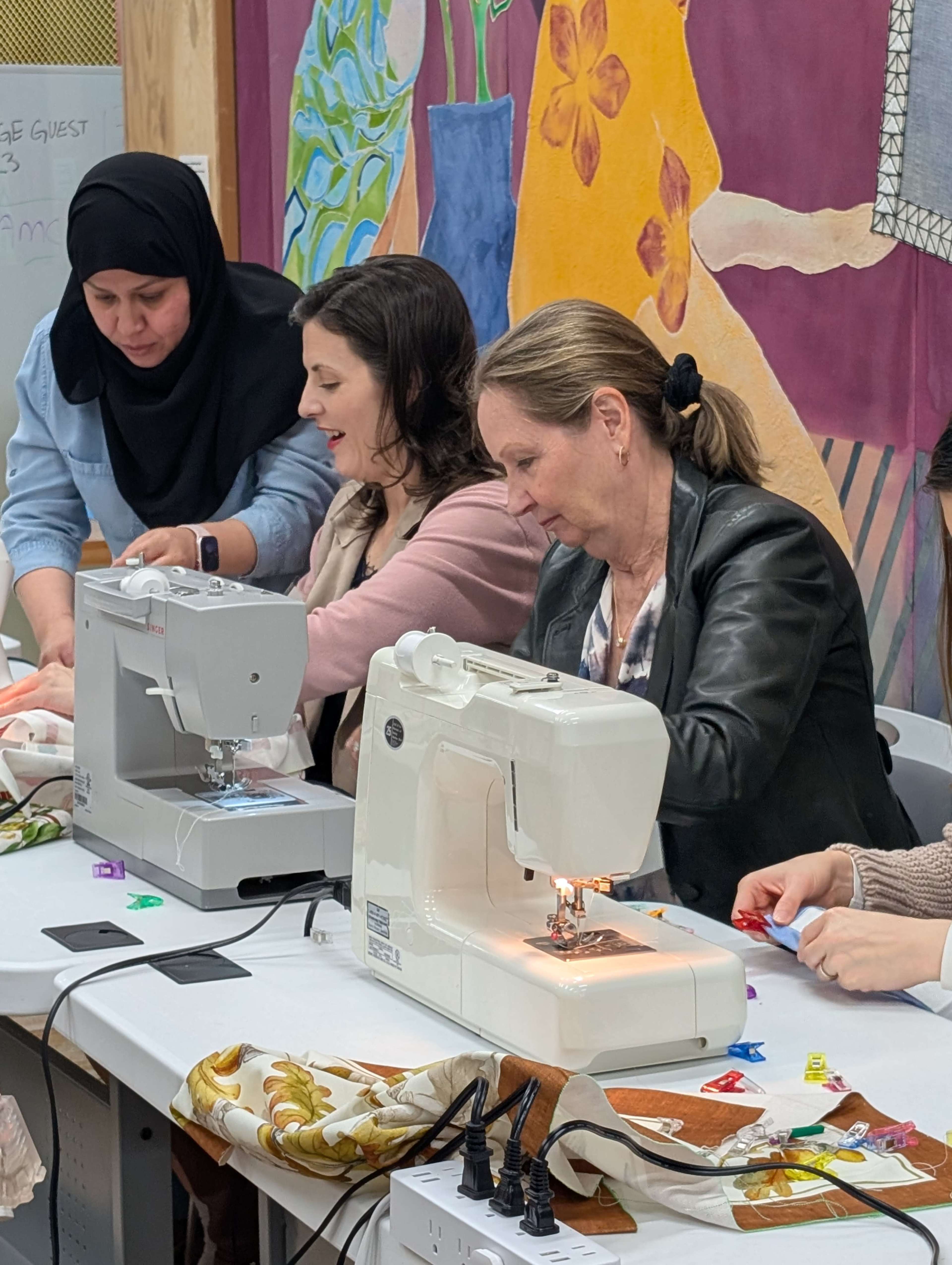 Three women are seated at a table, using sewing machines to work on fabric projects, with colorful artwork on the wall behind them.