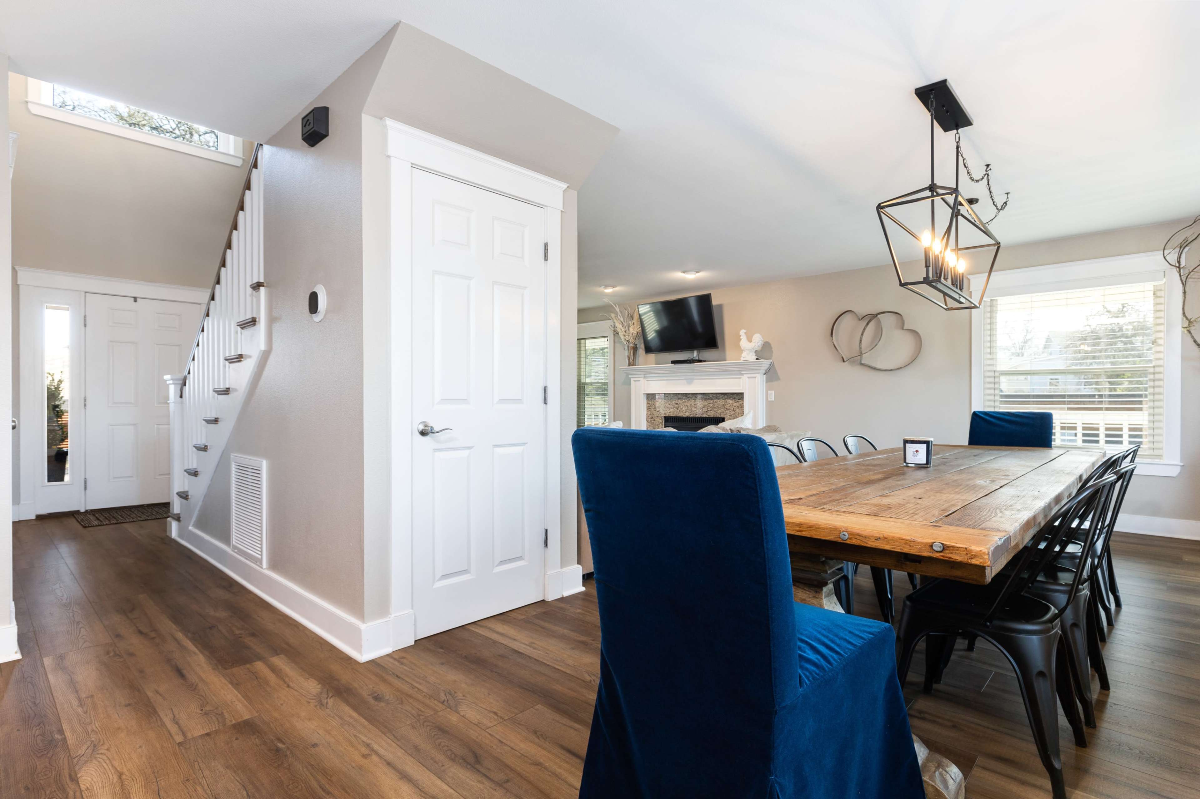 A modern dining area with a wooden table surrounded by blue chairs, leading to a staircase and an open living space with a fireplace and television.