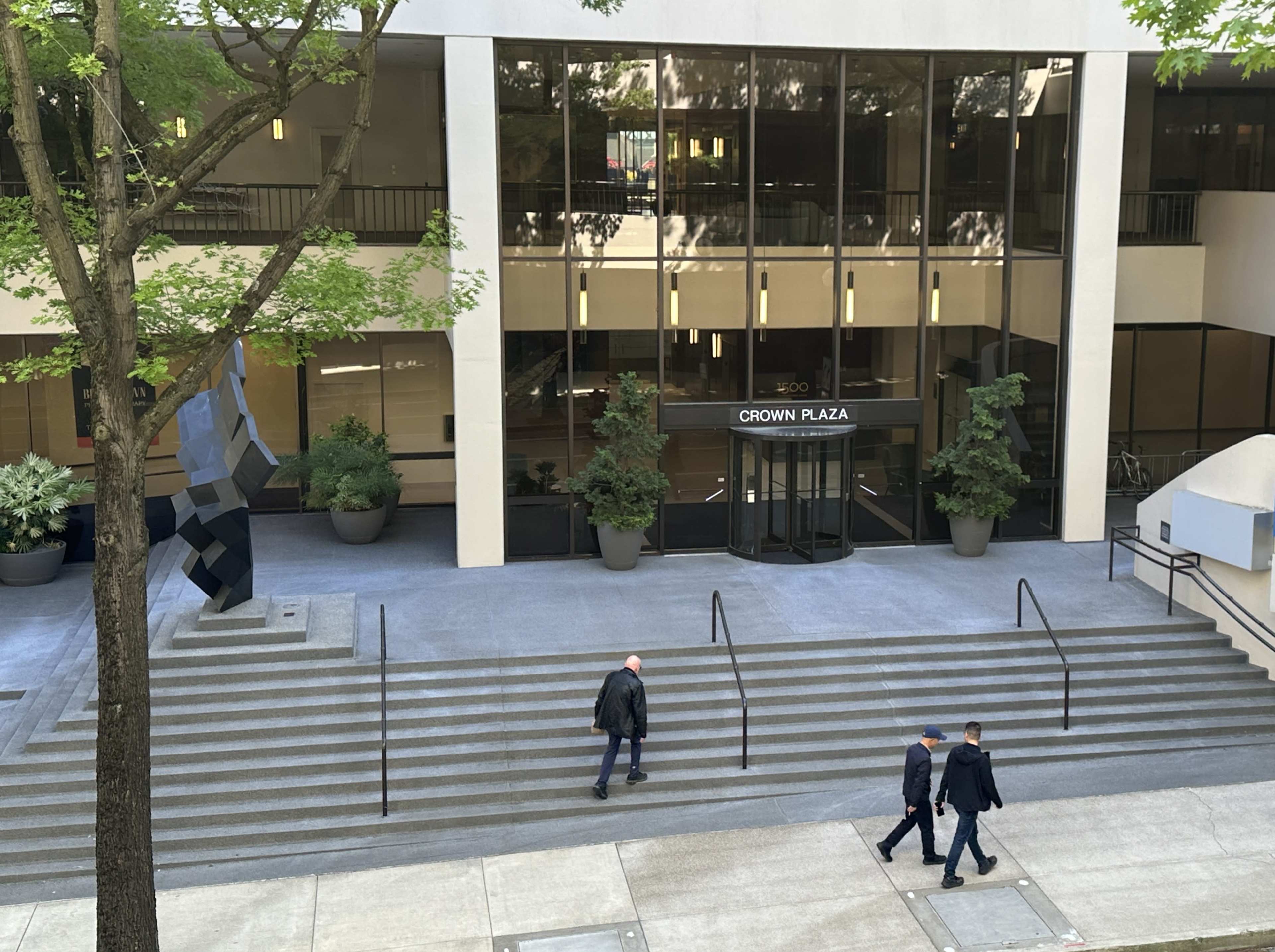 Two people walk down the stairs in front of the Crown Plaza hotel, flanked by large potted plants and a geometric sculpture.