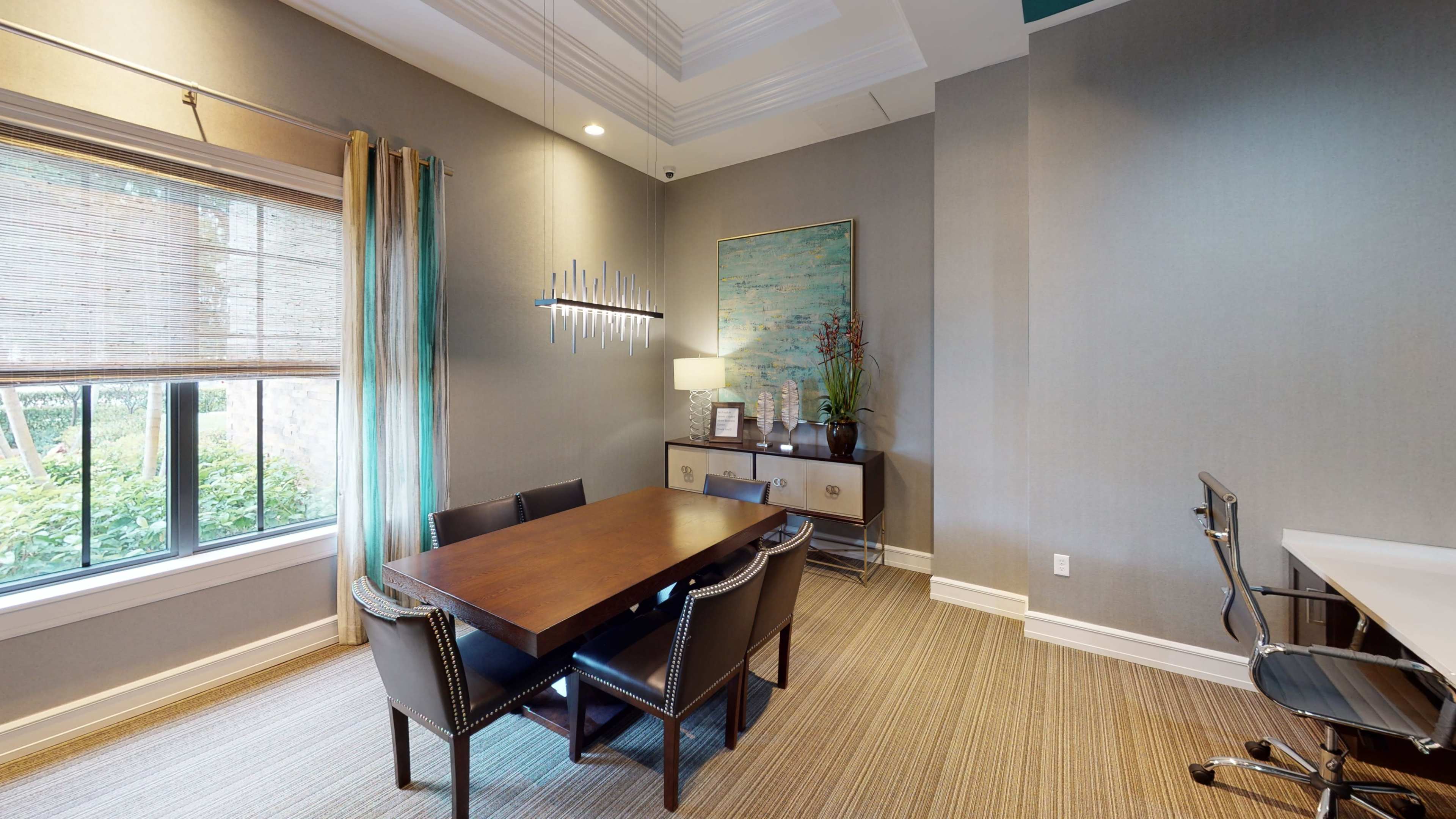 A well-lit office space featuring a wooden table surrounded by chairs, a console table with decor, and large windows with shades.