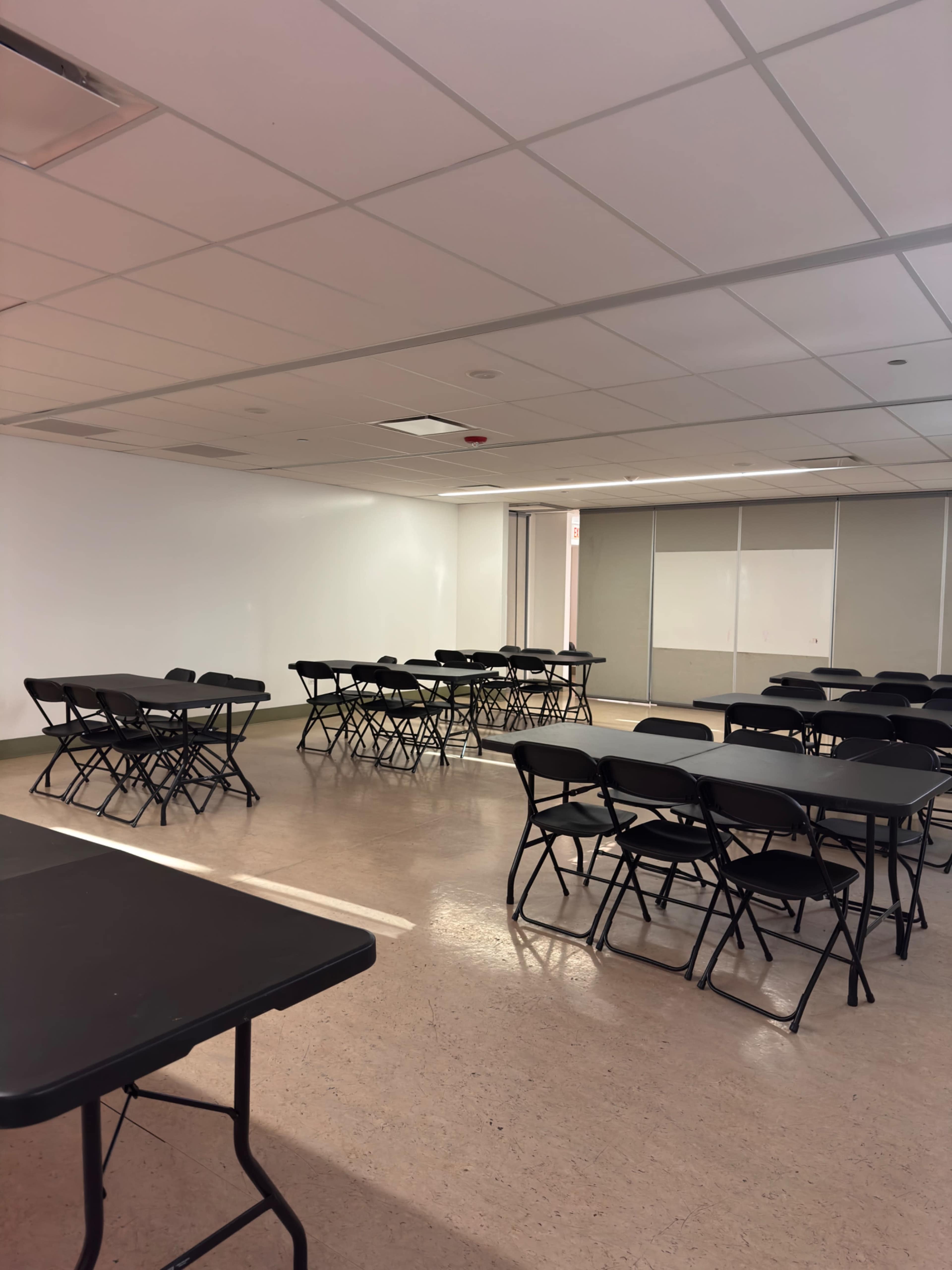 The image depicts a spacious room arranged with several black folding tables and chairs set up for an event or meeting.