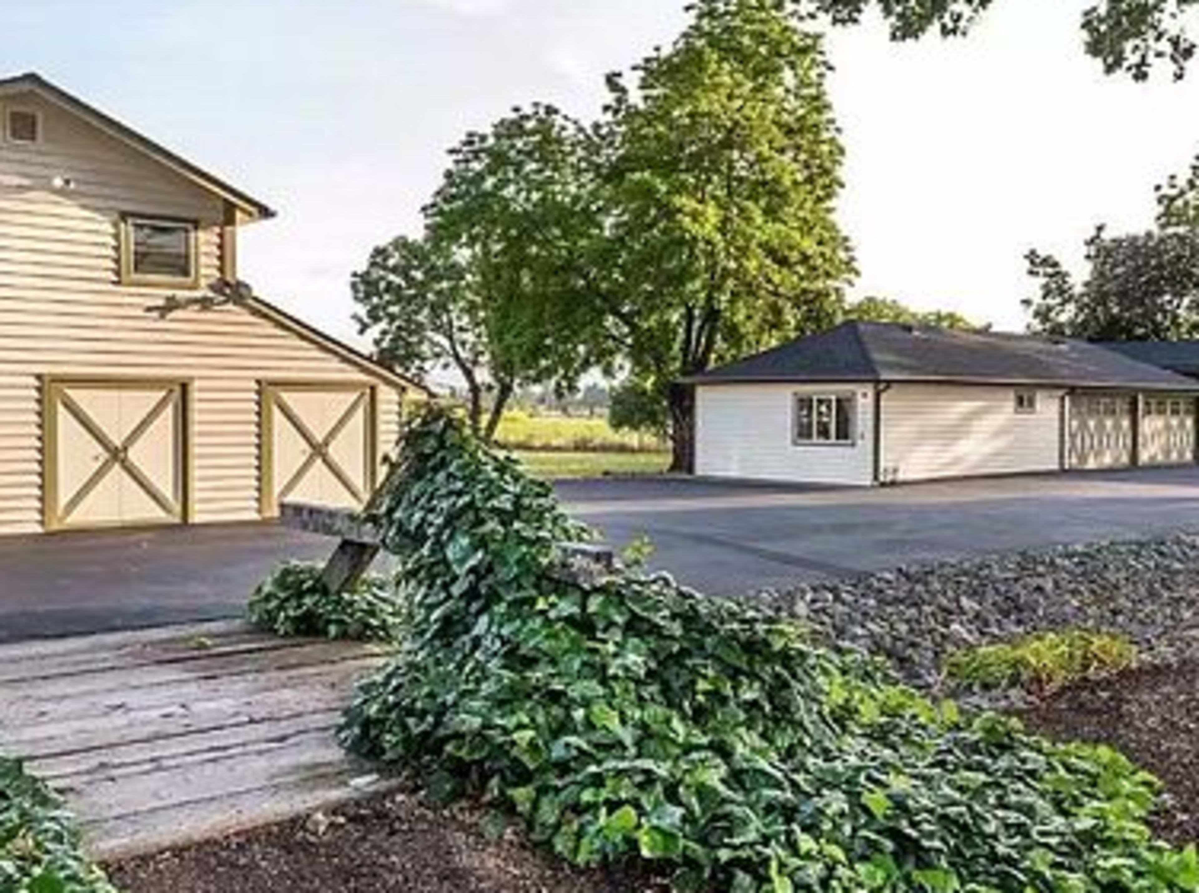 A light-colored barn and a separate building beside a paved driveway, with green vegetation in the foreground.