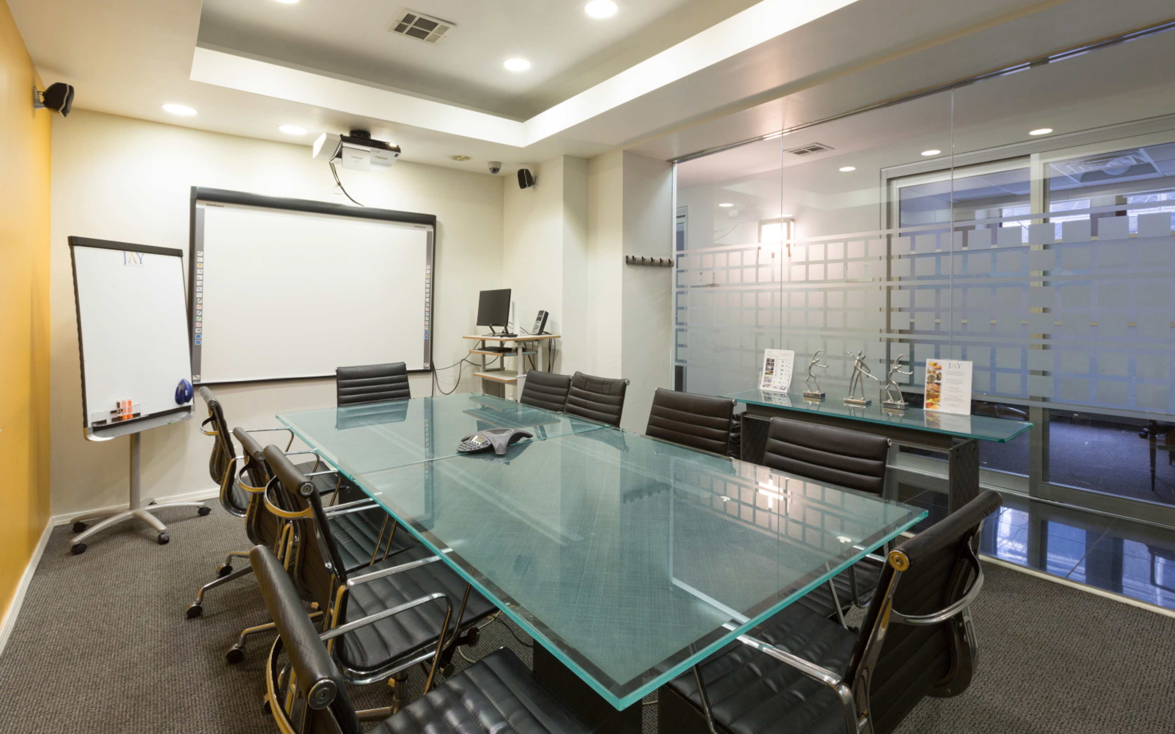 A modern conference room features a large glass table surrounded by black chairs, with a projector, whiteboard, and glass wall.