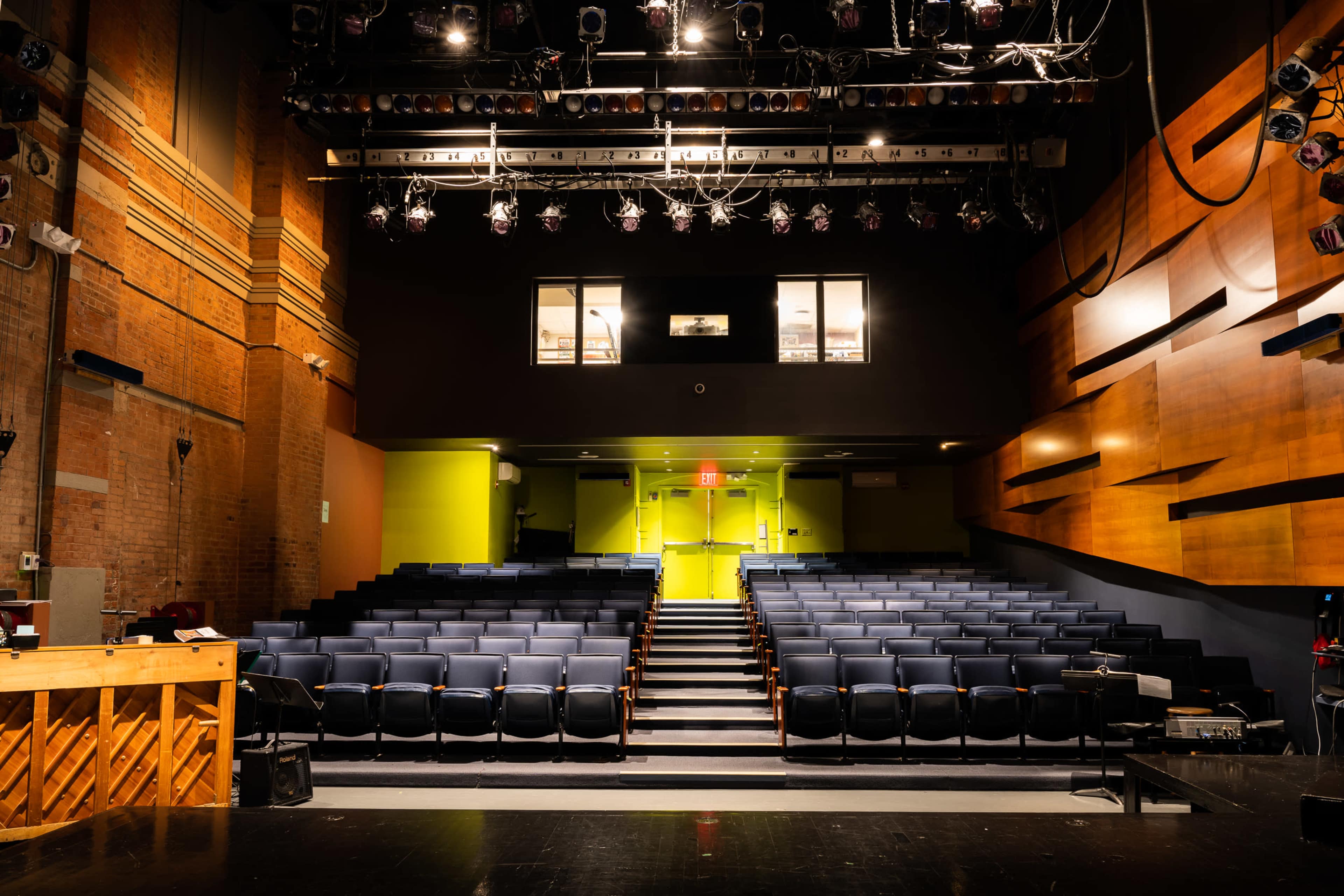 A theater interior with rows of blue seats facing a stage, illuminated by overhead lighting and featuring large windows above the entrance.