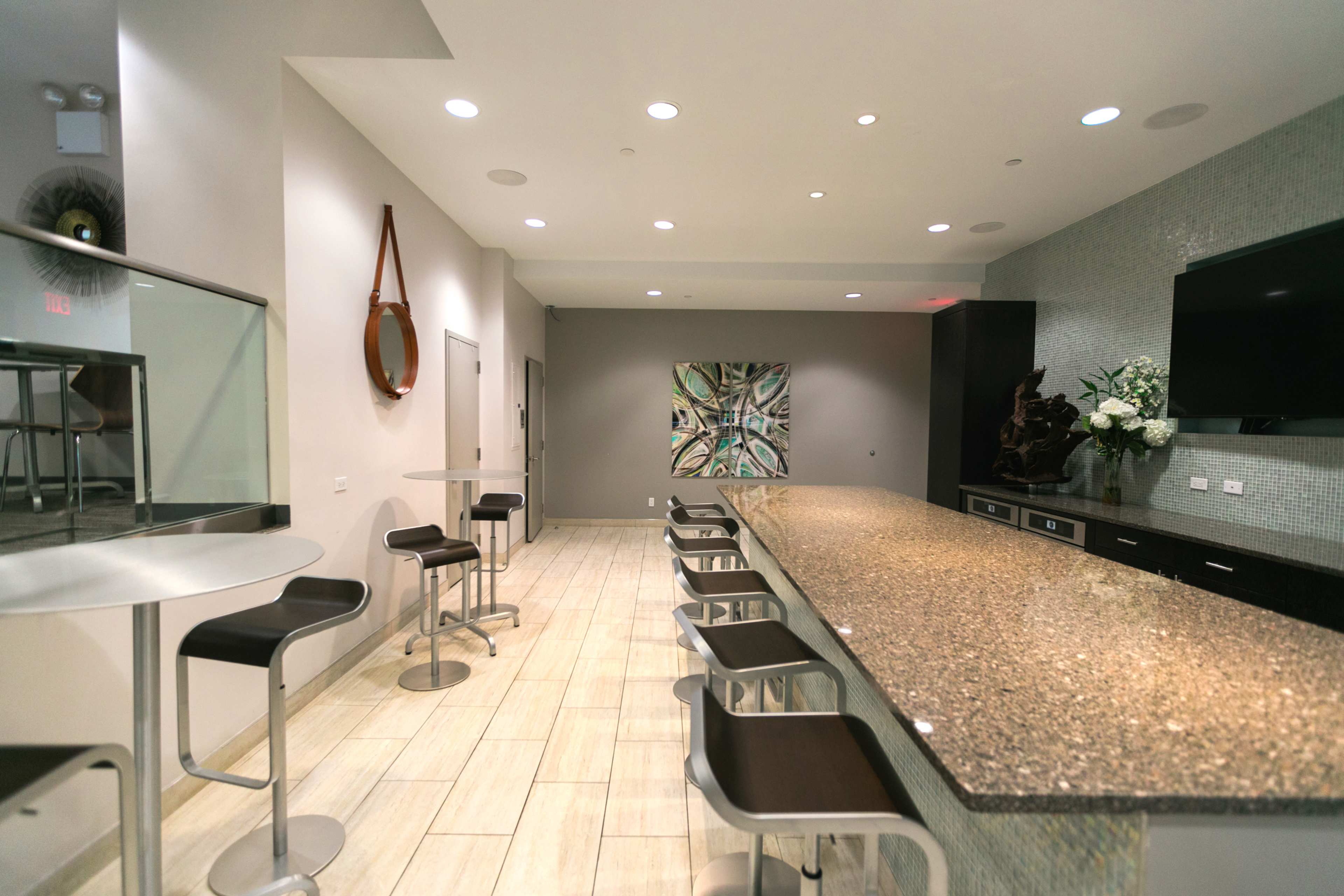 The image shows a modern, minimalistic bar area featuring a long granite countertop with several stools and a large wall-mounted TV, illuminated by overhead lighting.