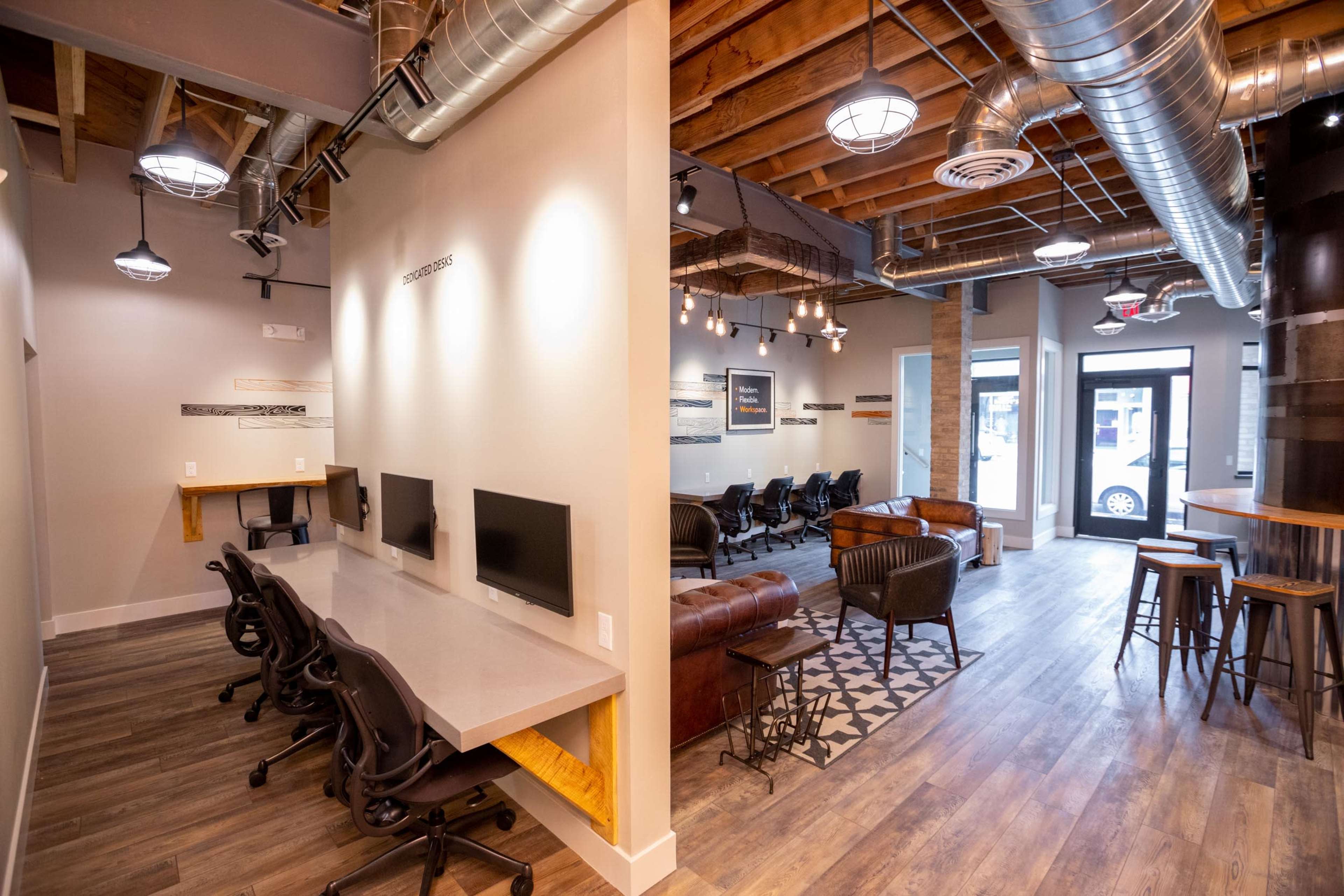 The image shows a modern office space featuring a workstation area on the left with multiple computers and a lounge area on the right with leather seating and decorative lighting.