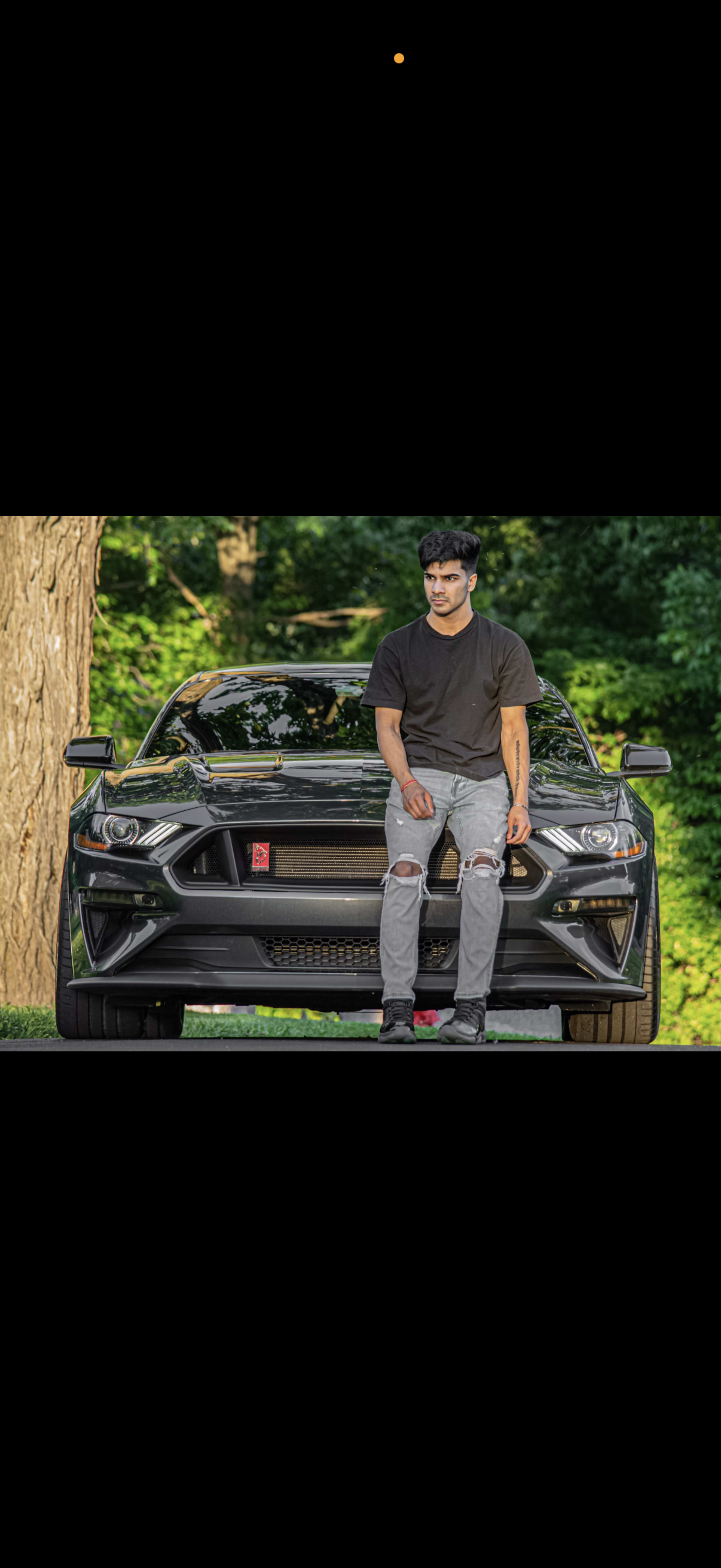A young man in a black t-shirt and ripped jeans sits on the hood of a black sports car parked in a wooded area.
