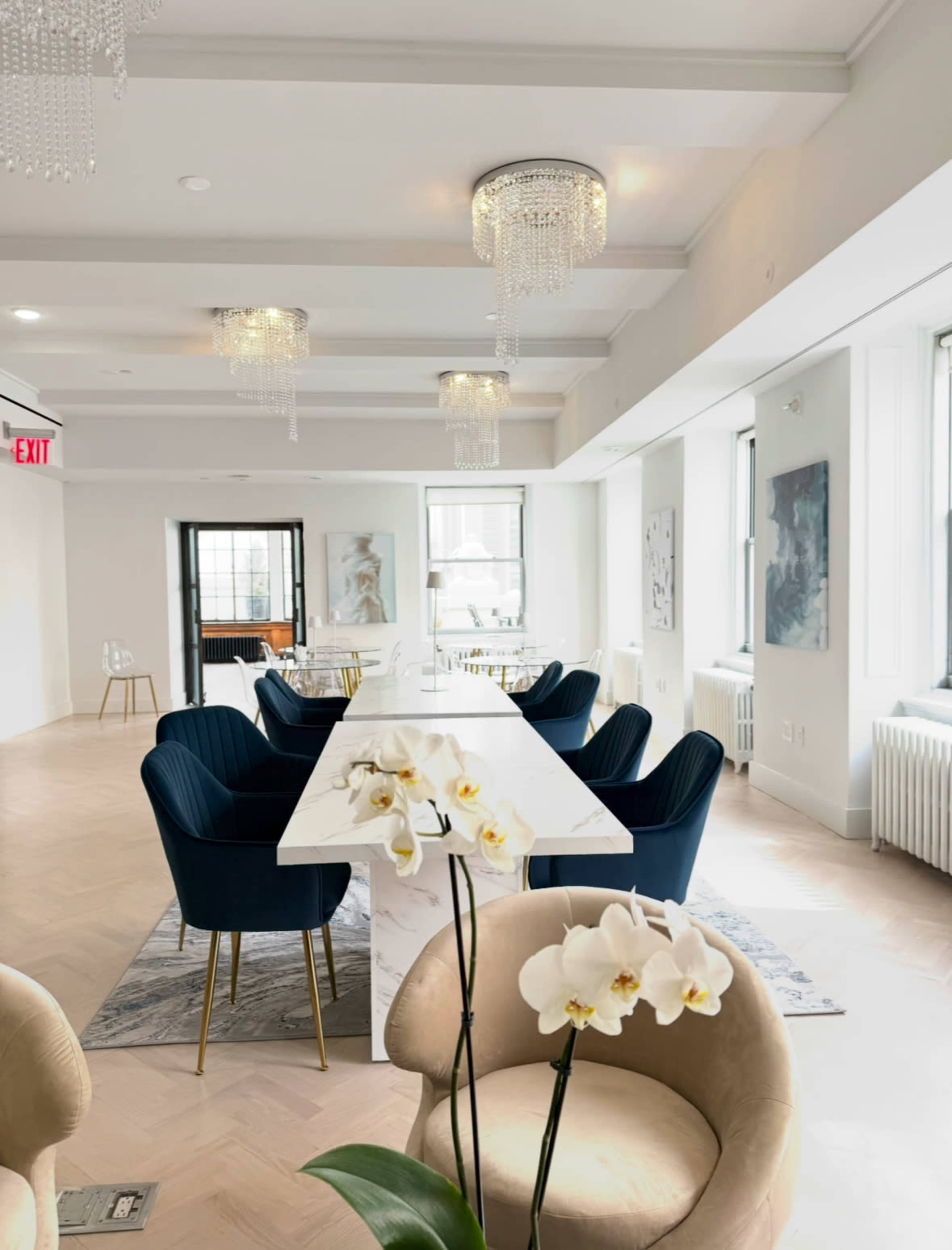The image shows a modern, light-filled dining or meeting area featuring a long white table, blue chairs, and decorative chandeliers.