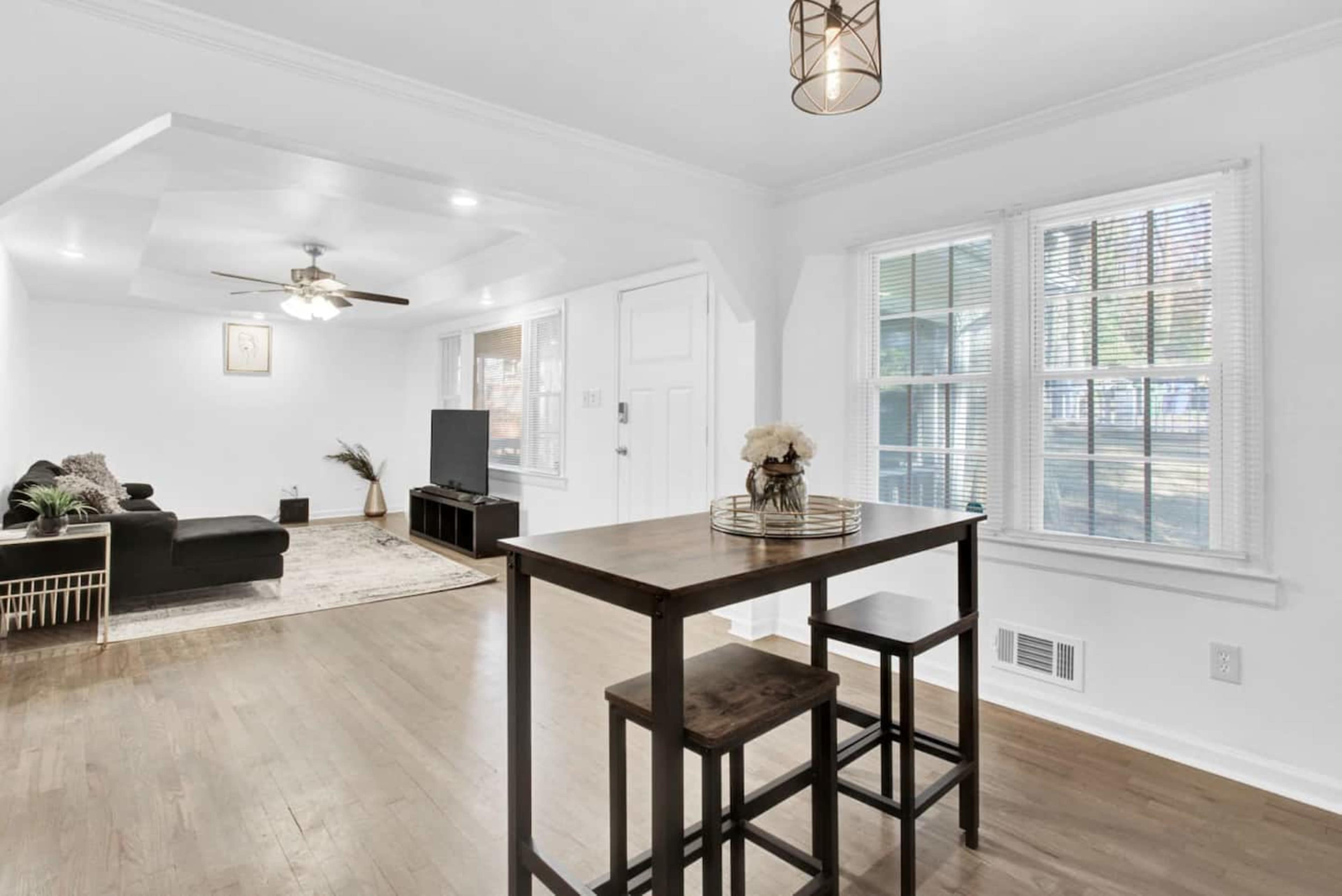 A well-lit living space with a dining table and chairs, a sofa, a television, and large windows.