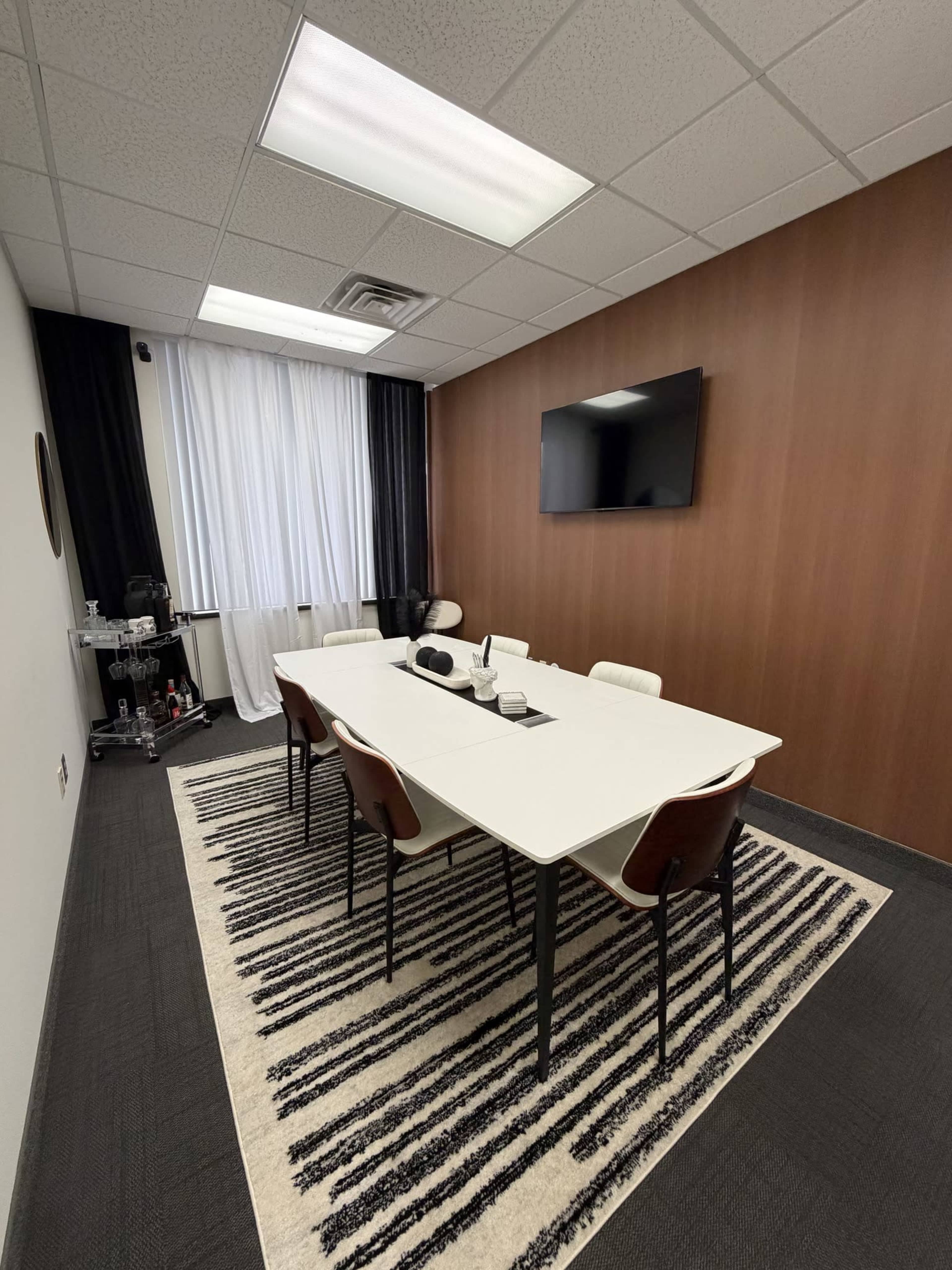 The image shows a conference room with a long white table, several chairs around it, and a television mounted on a wooden wall.