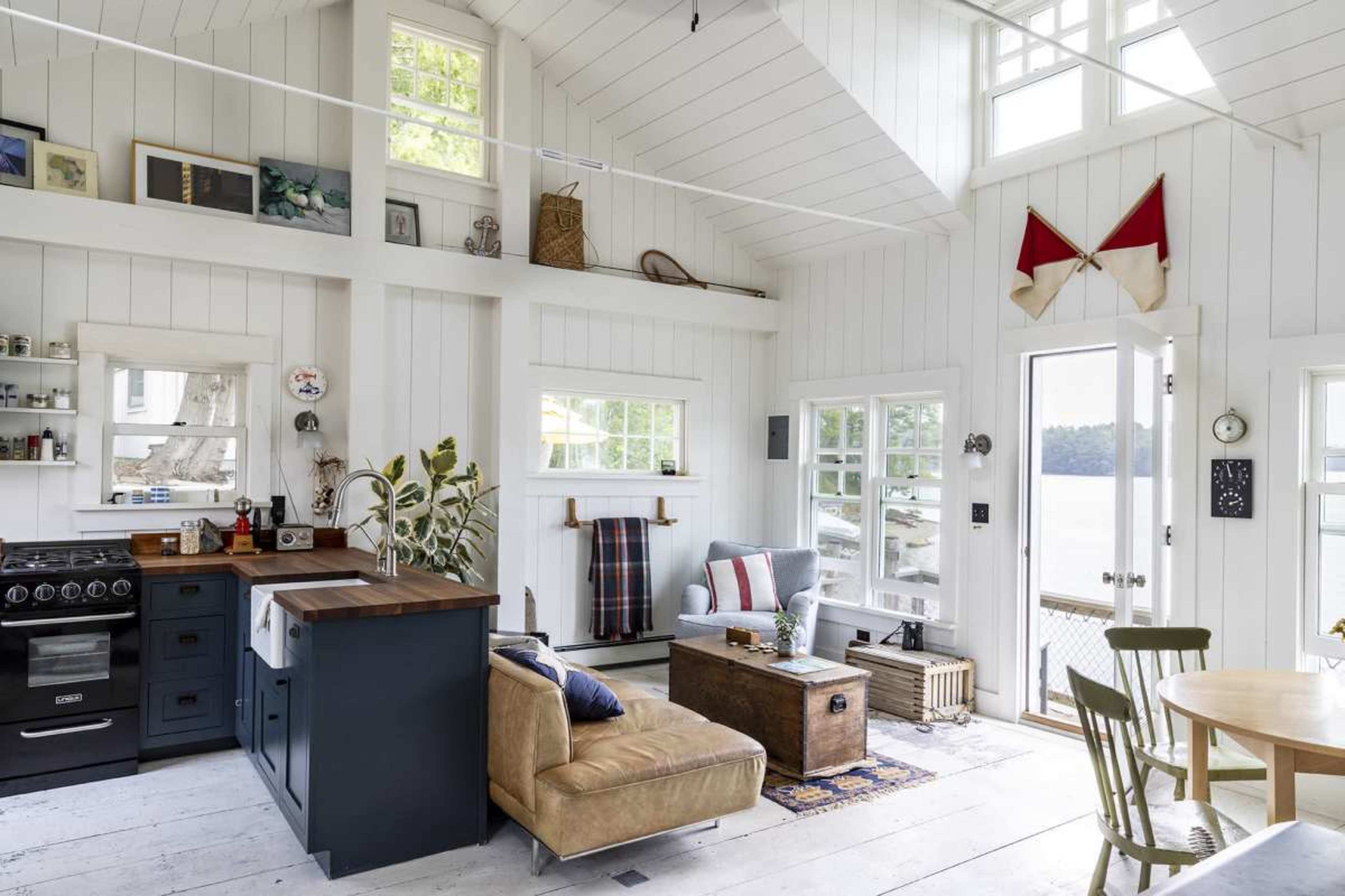 The image shows a bright, modern interior of a small cottage, featuring a kitchen area with a black stove, a living space with a couch and coffee table, and large windows offering a view of a water body.