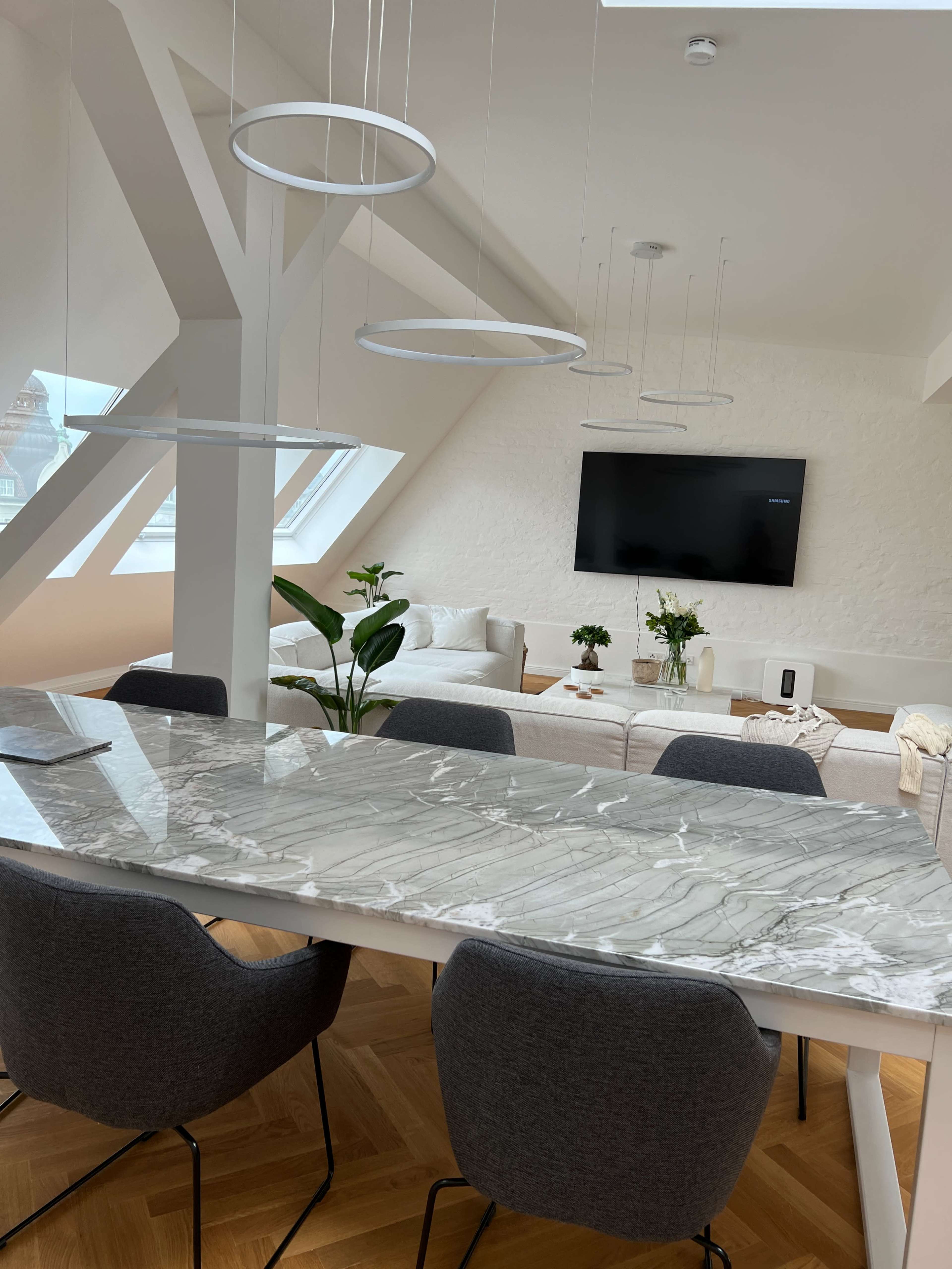 A modern dining area features a marble table surrounded by gray chairs, with a spacious living room visible in the background.