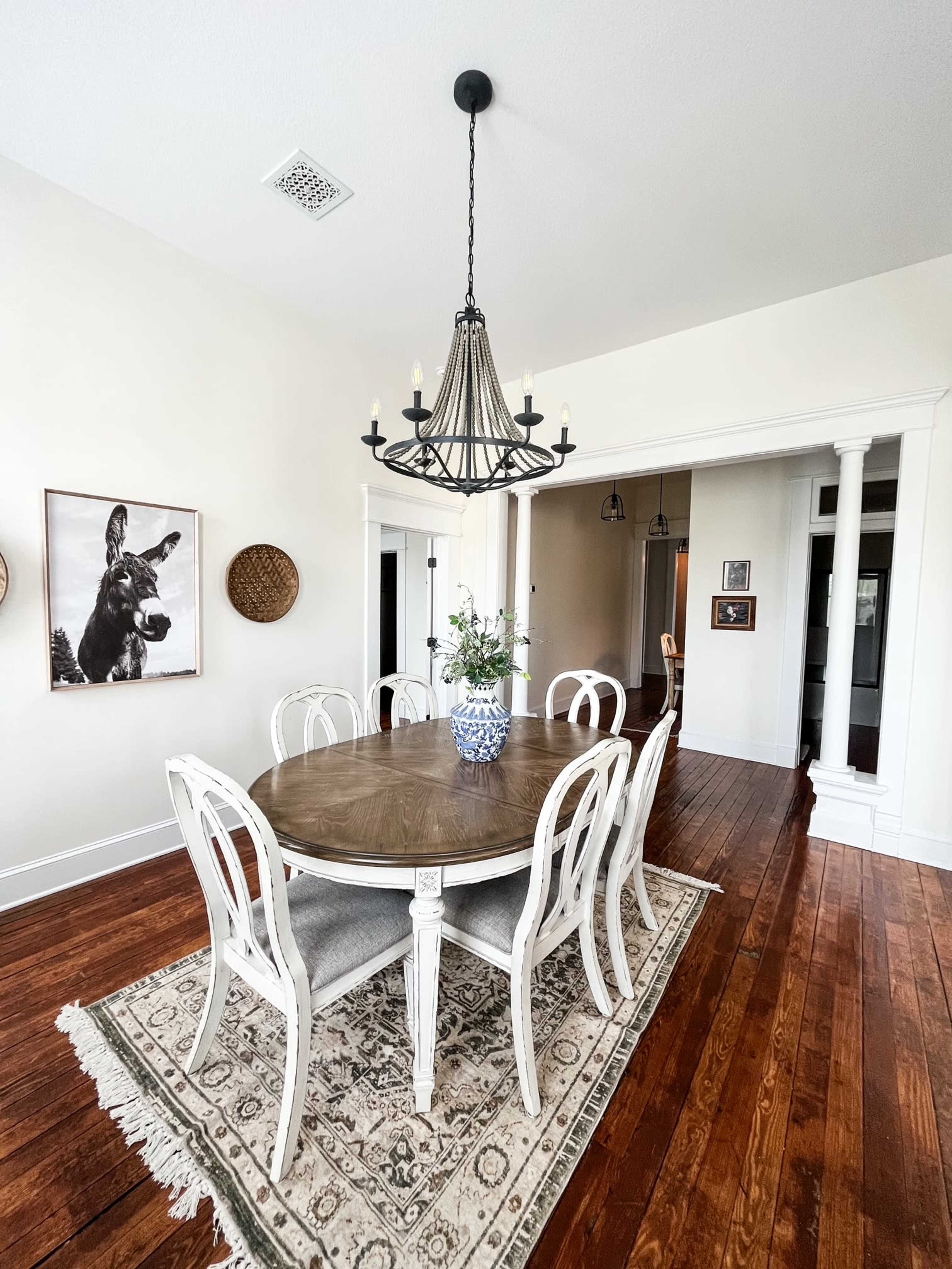 A dining room features a wooden table surrounded by six white chairs, centered on a patterned rug, with a chandelier overhead and decorative wall art.