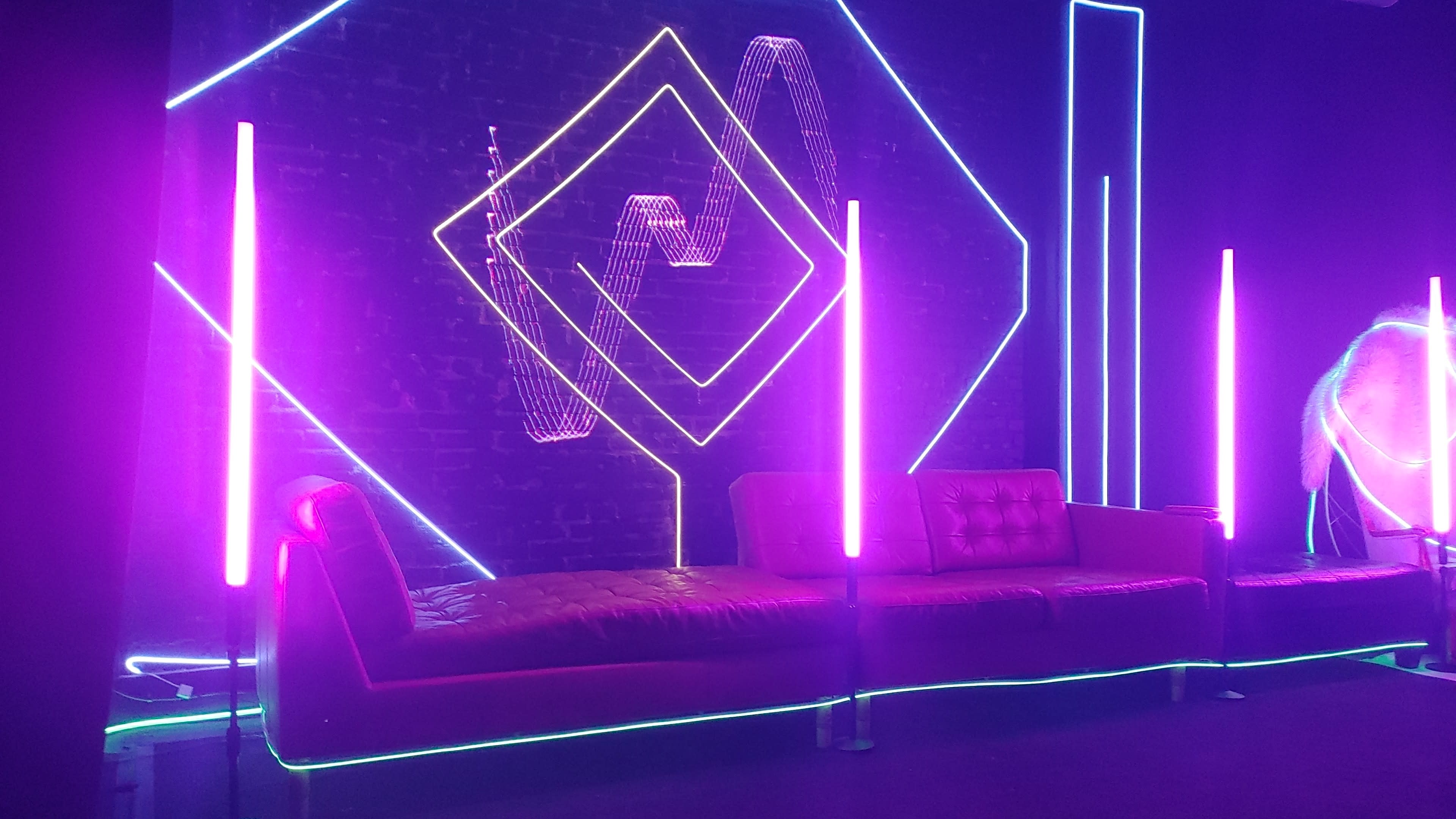 The image features a modern lounge area with a red couch and glowing neon light installations in purple and blue against a black brick wall.