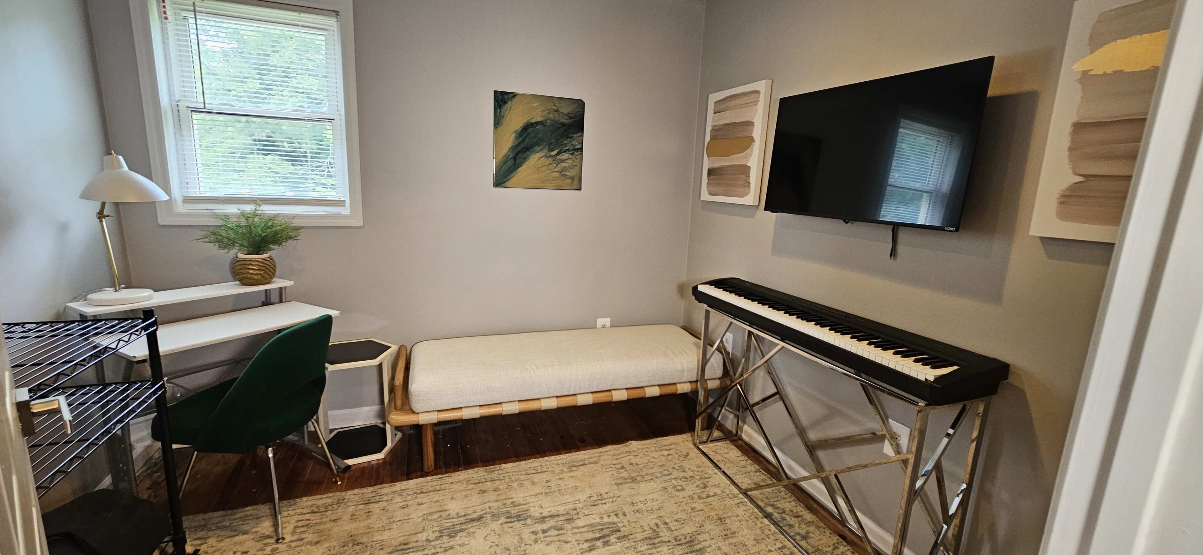 A small, well-lit room features a desk with a lamp, a green chair, a keyboard on a stand, a bench, a television mounted on the wall, and abstract art.