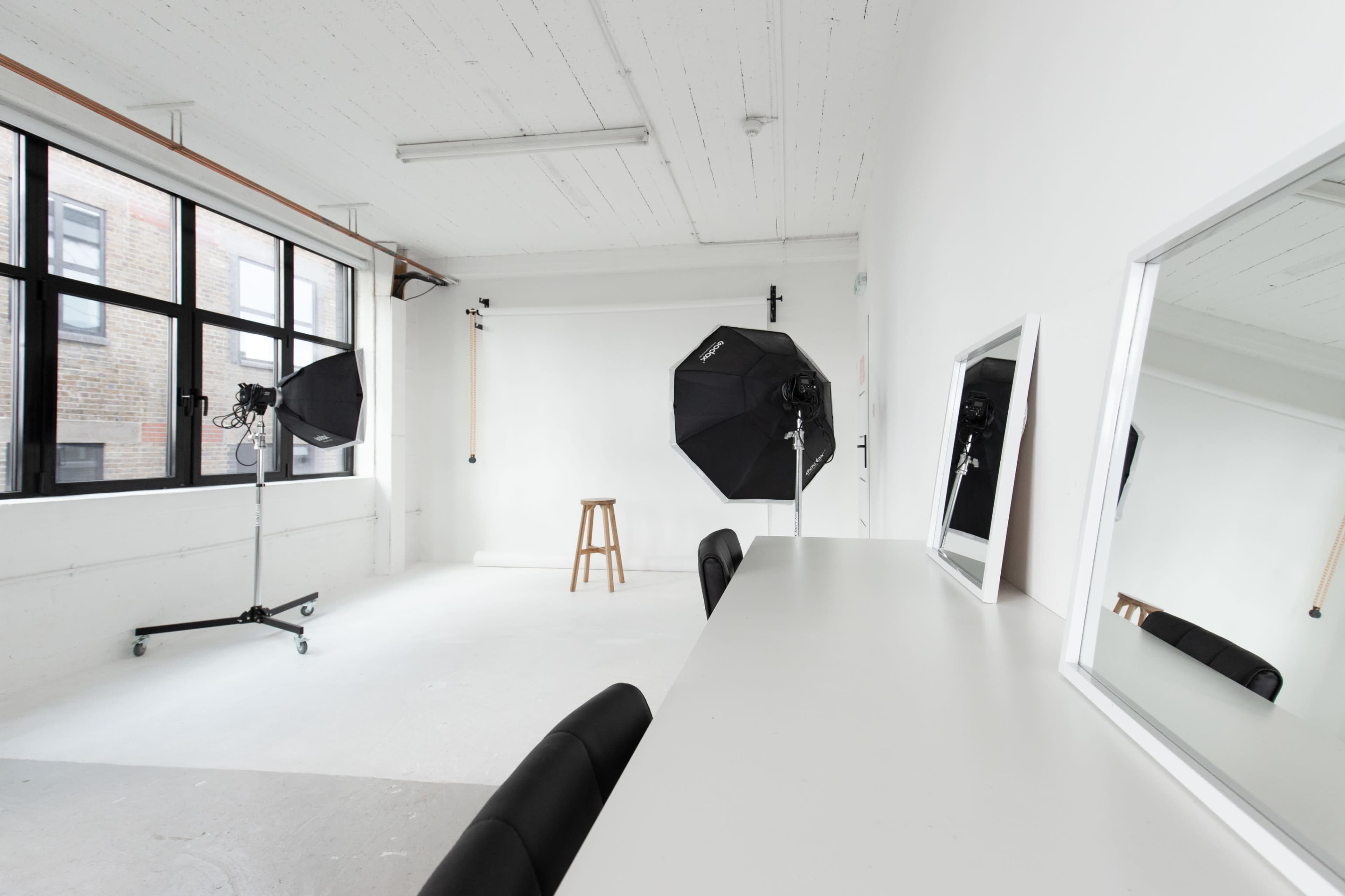 A spacious photography studio with a wooden stool, two softbox lights, and a white backdrop, along with a long table featuring two mirrors.