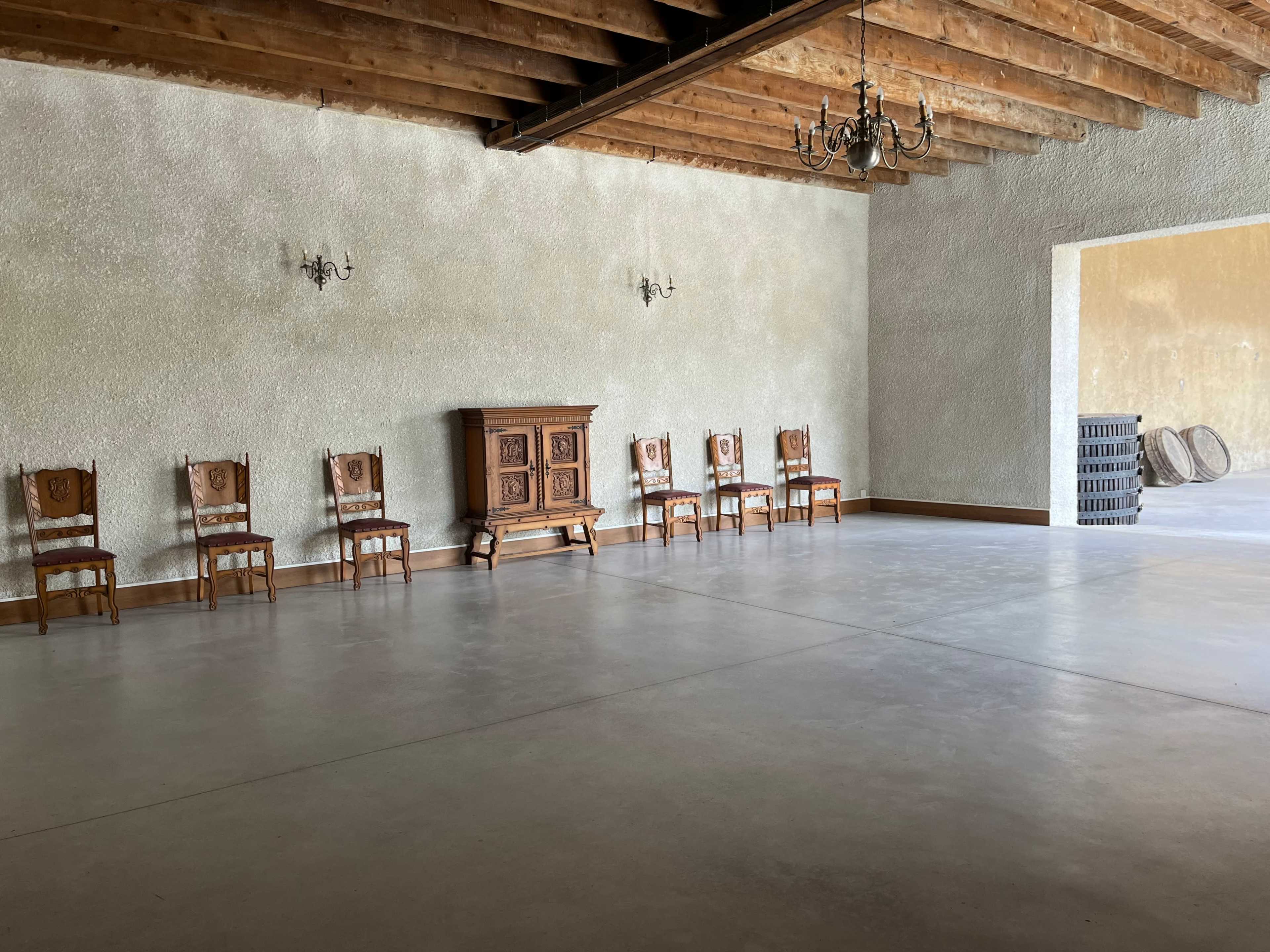 The spacious room features a row of wooden chairs against a textured wall, with a vintage cabinet and a chandelier hanging from the exposed beam ceiling.