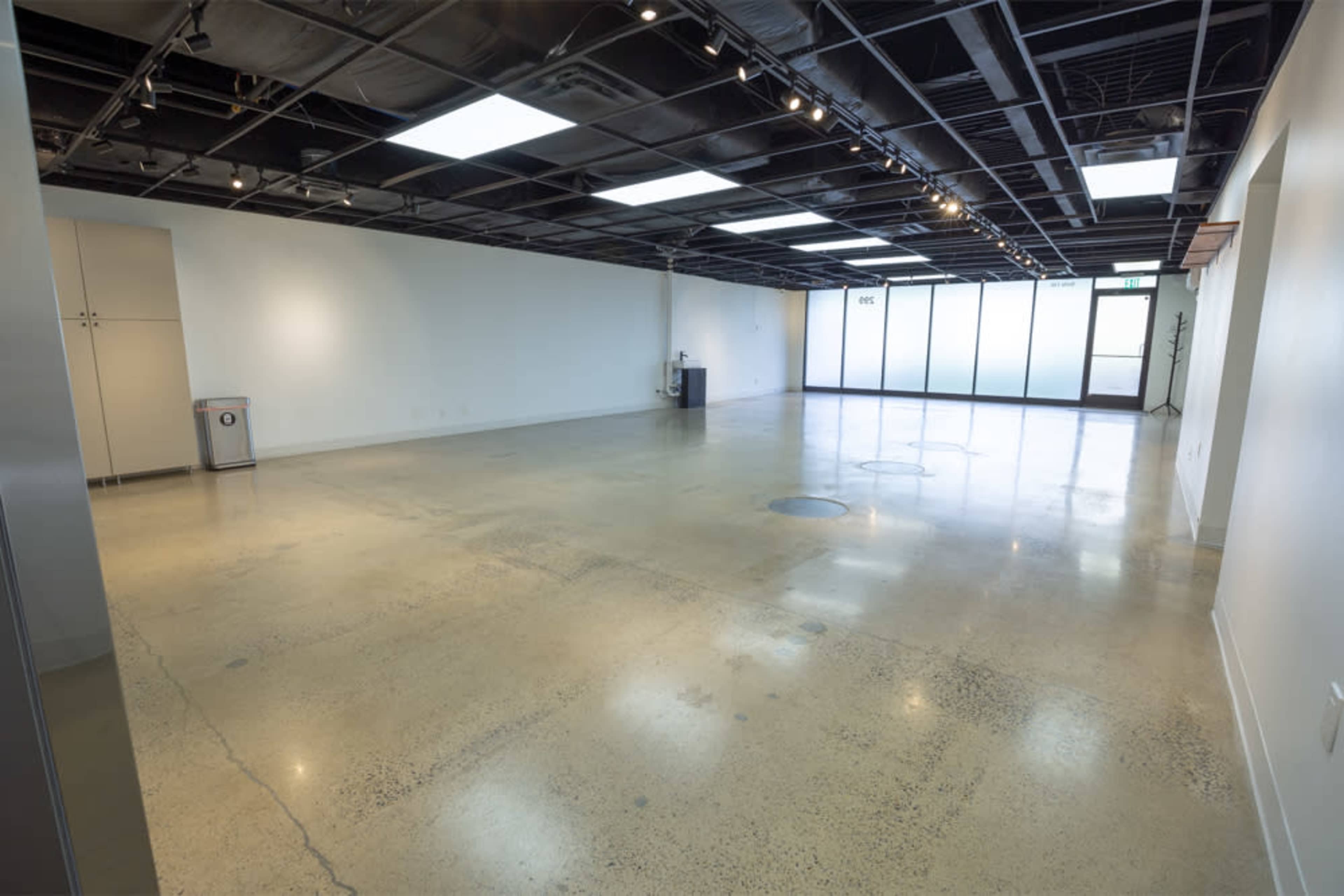 The image shows a spacious, empty room with polished concrete floors and overhead lighting, featuring large windows along one wall.