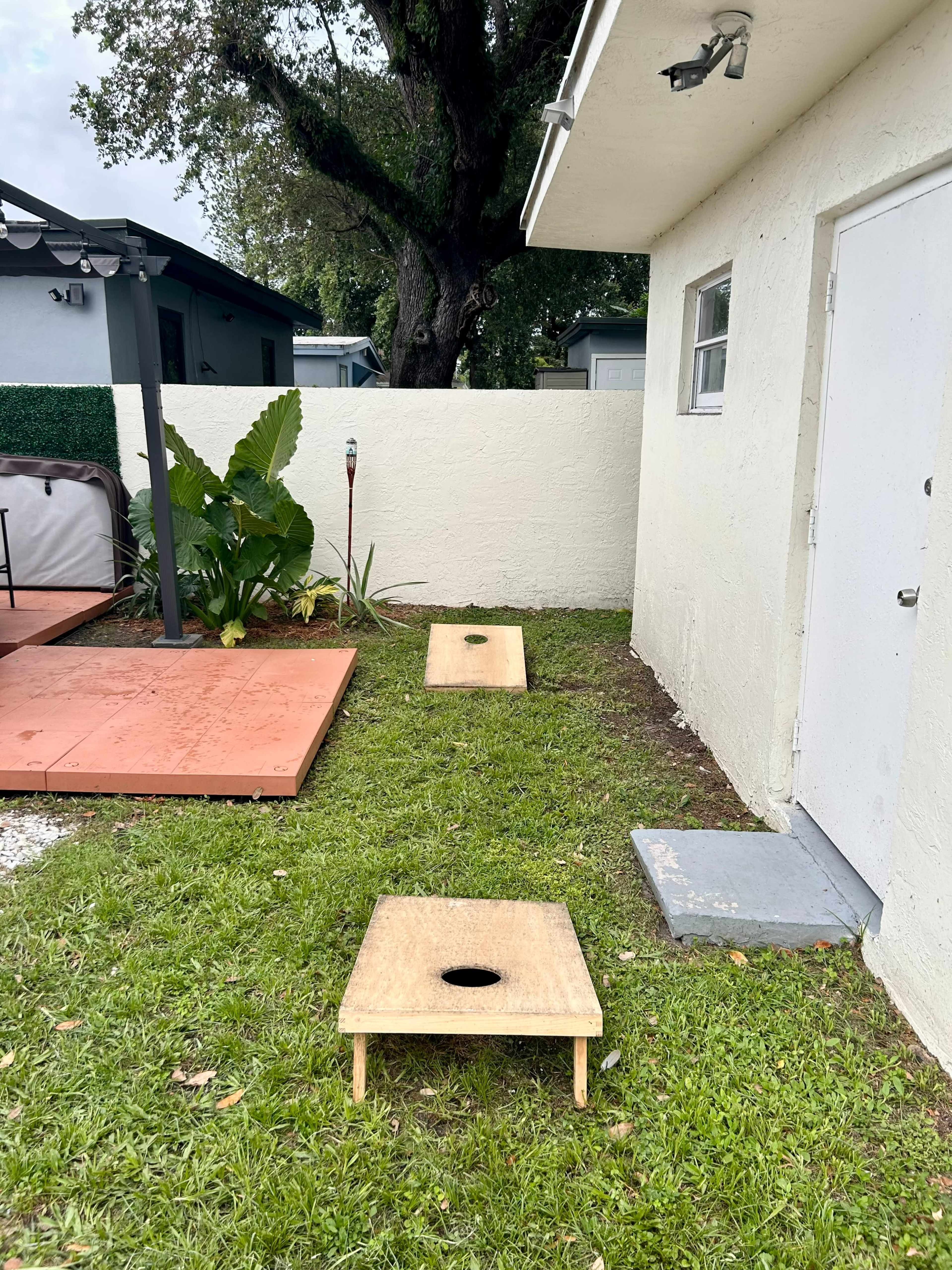 The image shows two wooden cornhole boards positioned on grass near a house, with landscaping and a wall in the background.