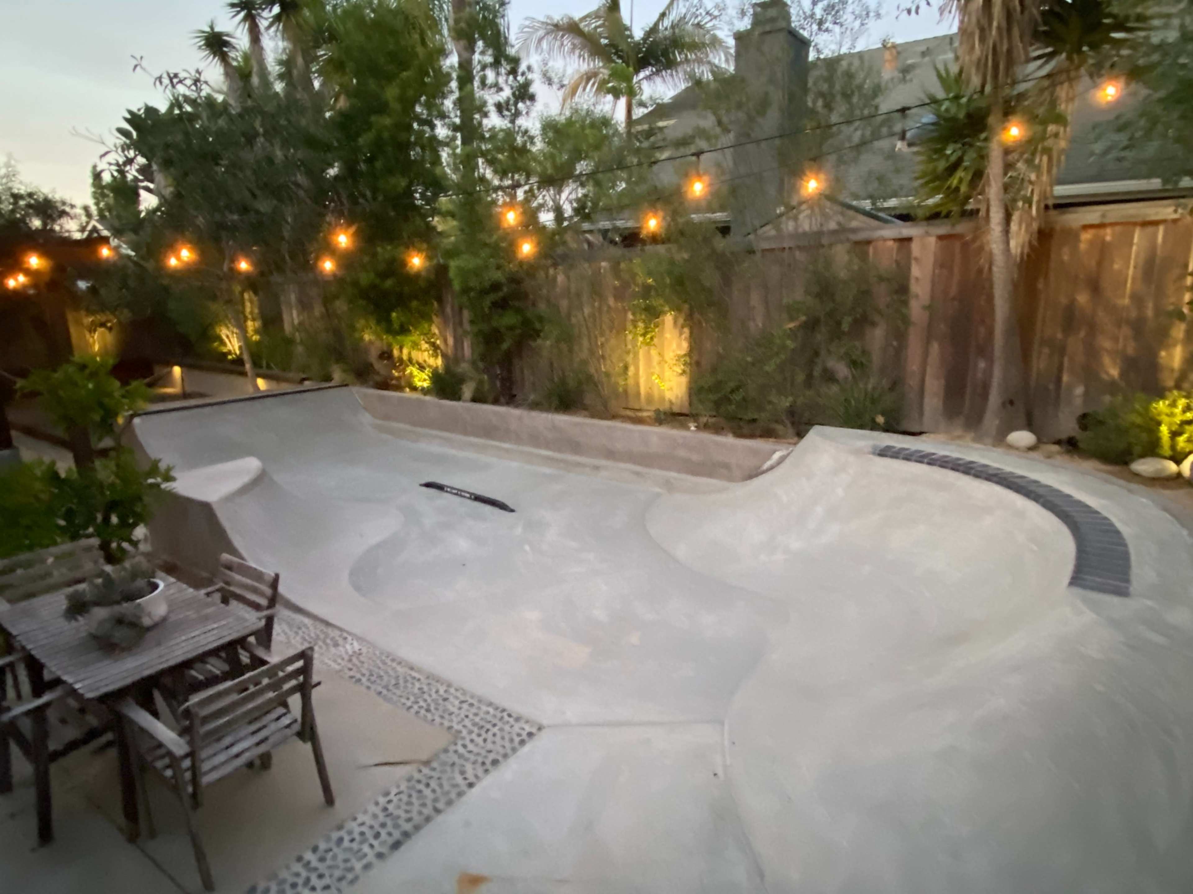 The image shows a modern backyard with a concrete skate bowl, surrounded by greenery and string lights illuminating the space.