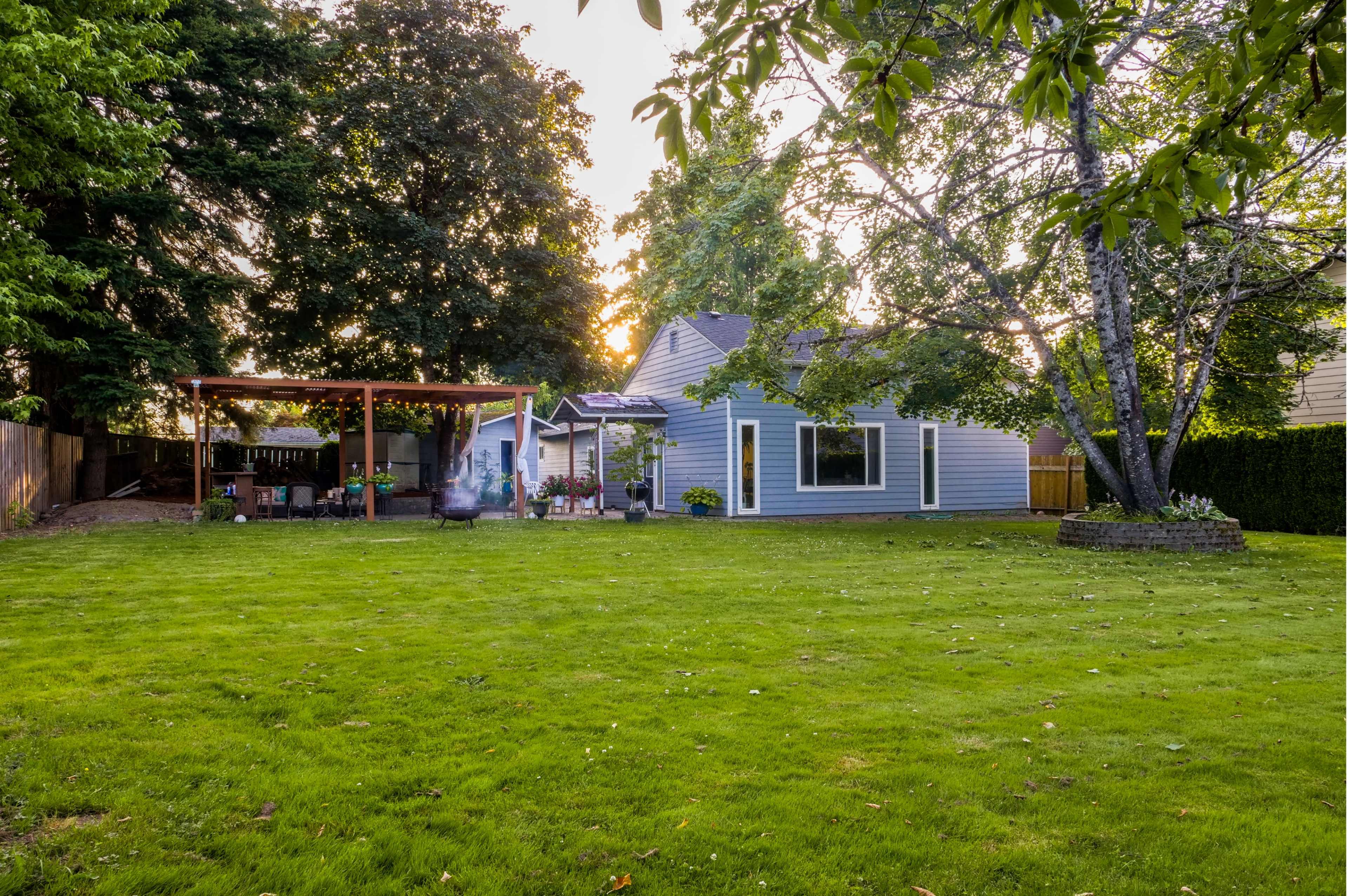 A spacious backyard features a green lawn, a wooden pergola, and a blue house with large windows.