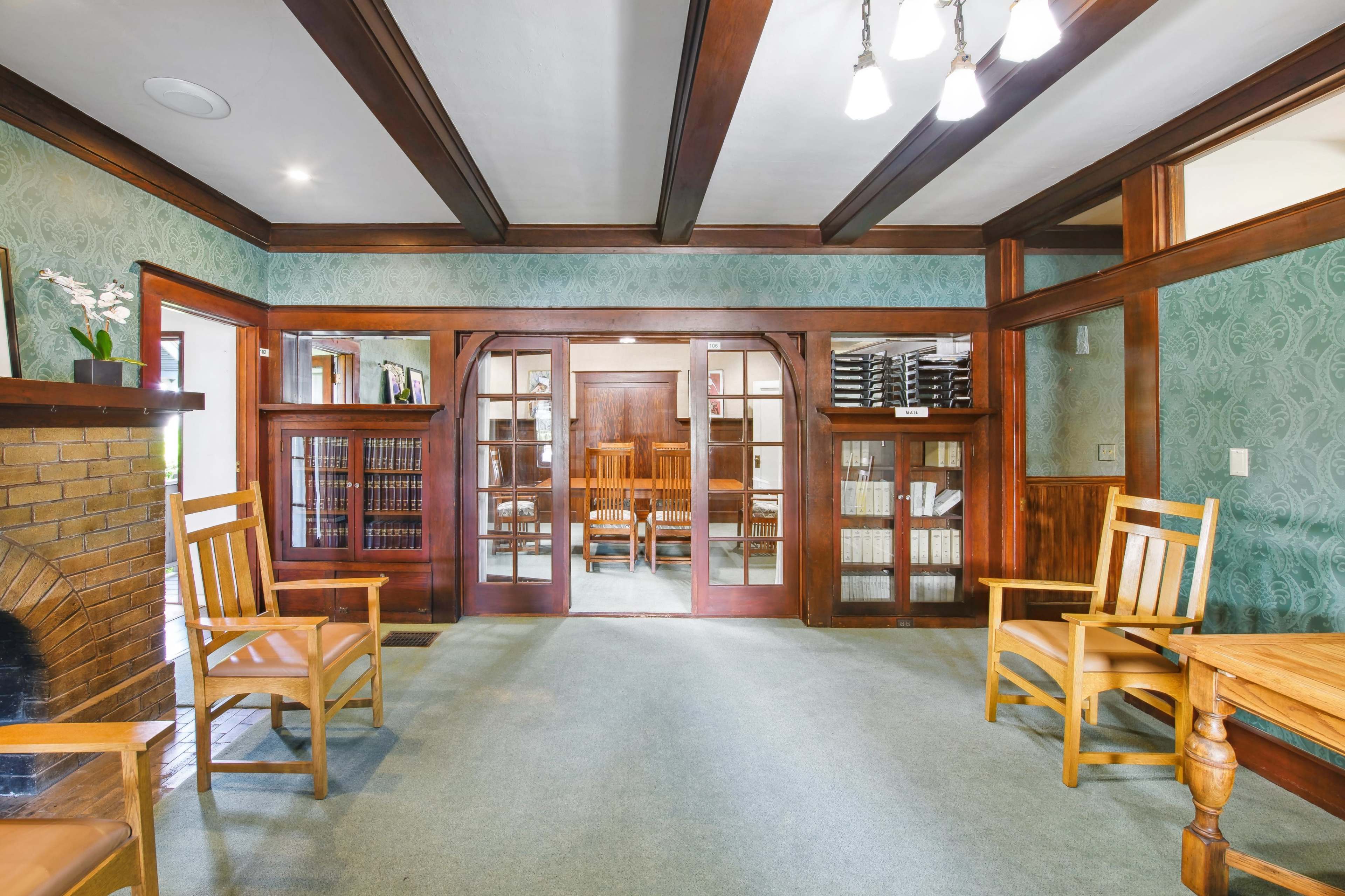 A room with wooden chairs and a dining area visible through glass doors, featuring patterned wallpaper and wooden beams on the ceiling.
