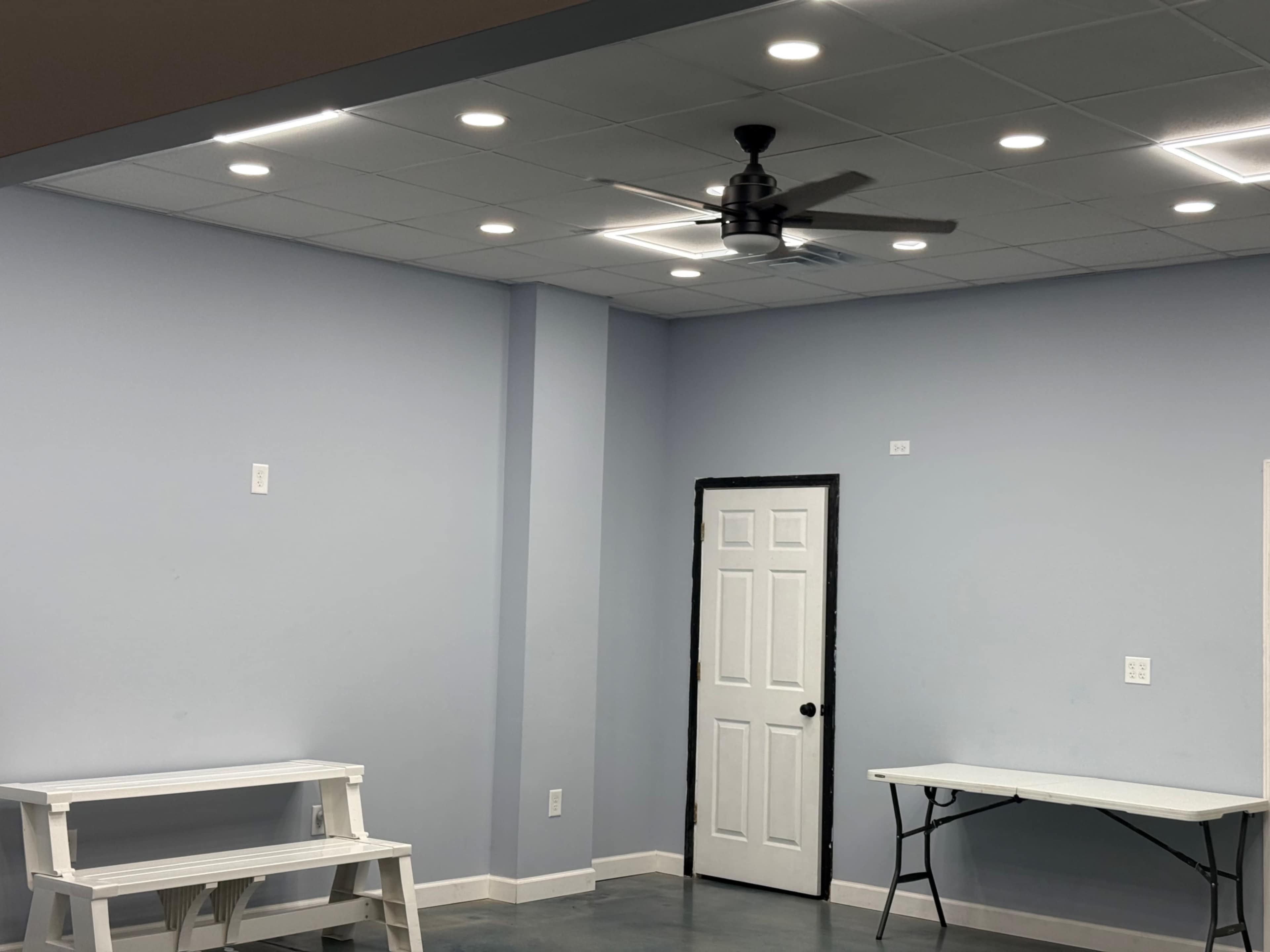 The image shows a partially furnished room with light blue walls, a ceiling fan, and two tables, one white and one folded, along with a closed door in the background.