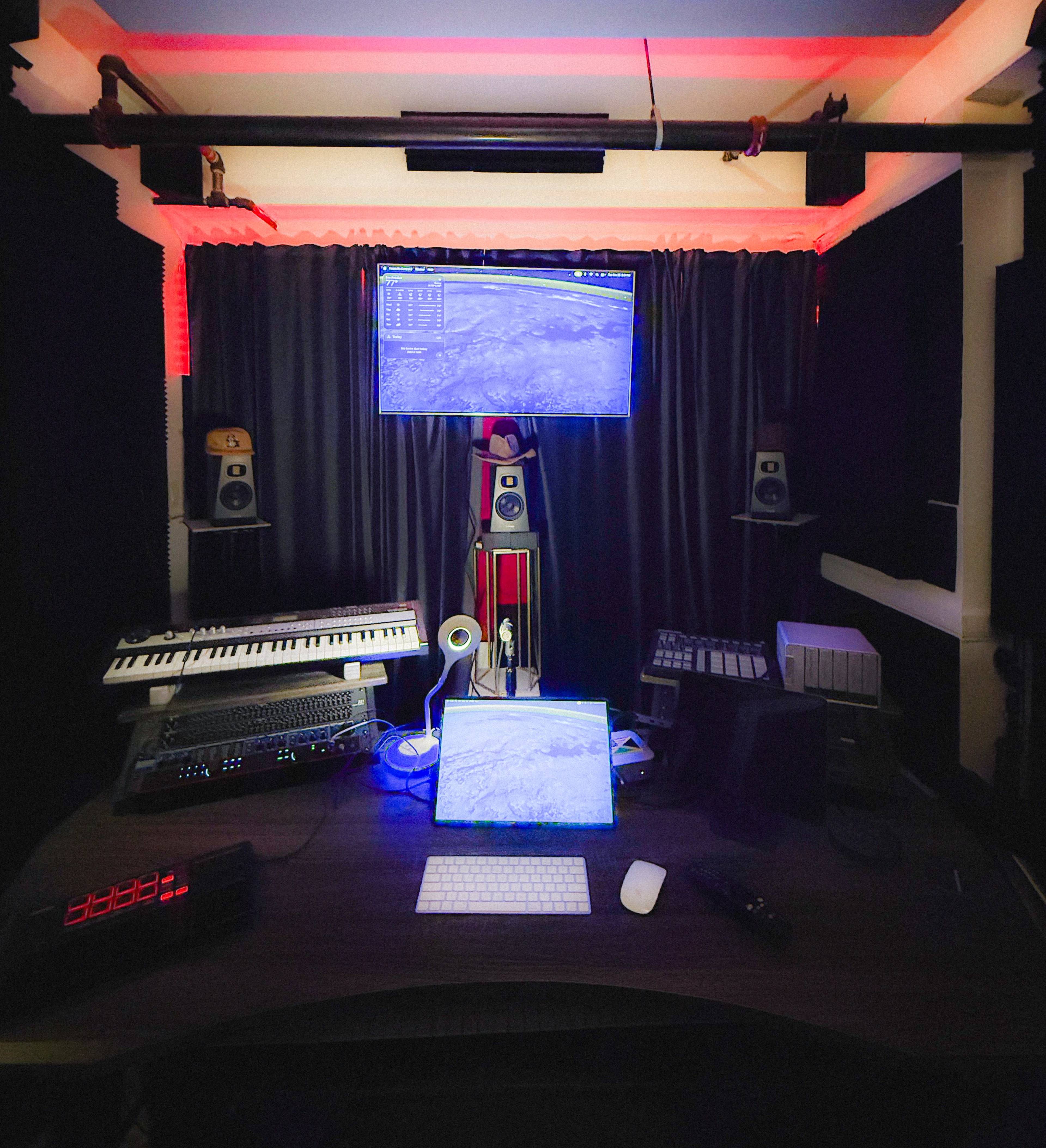 The image shows a music production studio with a desk featuring a computer, keyboard, and various music equipment, all surrounded by dark curtains and illuminated by red lights.