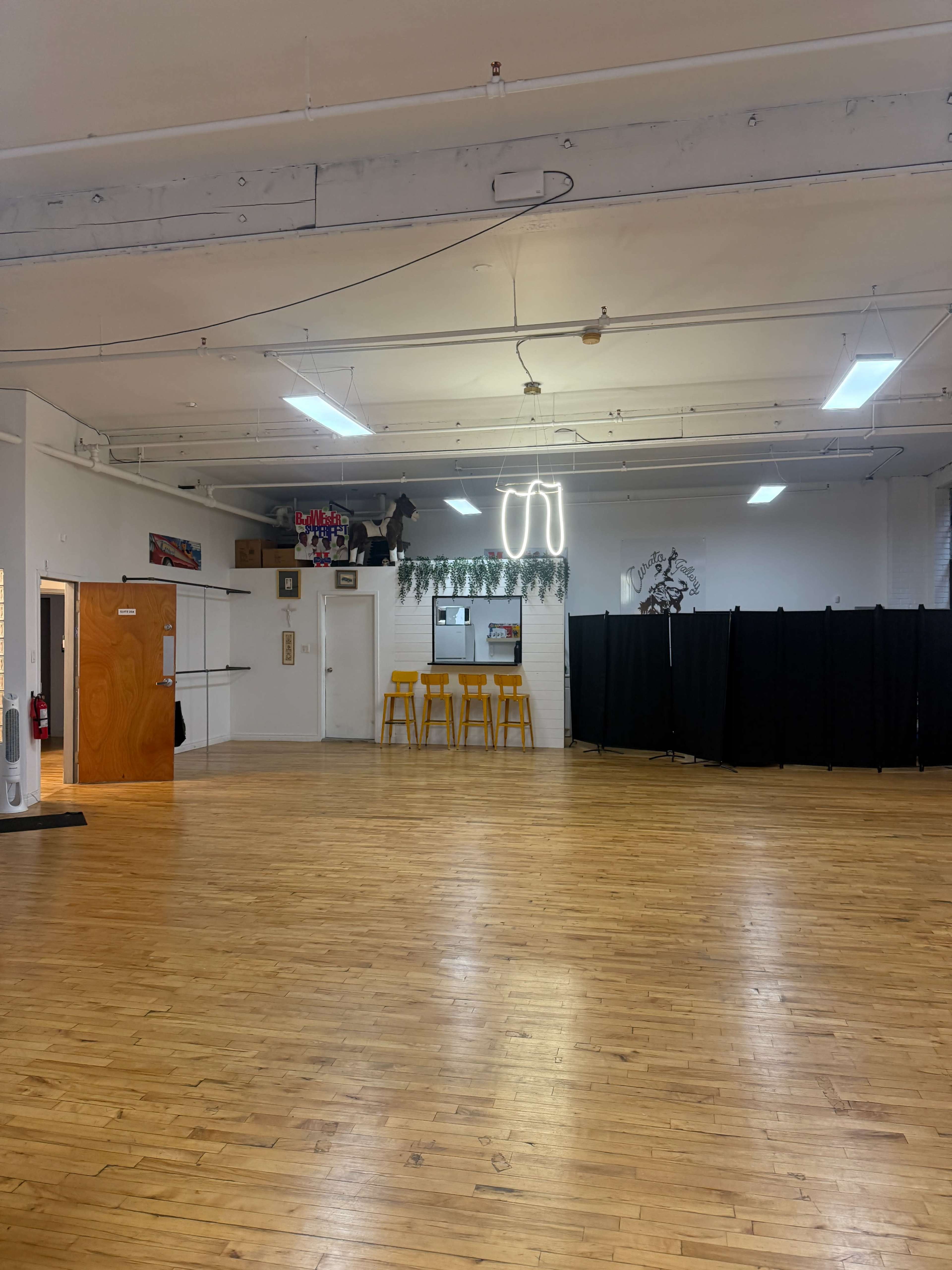 The image shows a wooden-floored studio space with a bar area featuring yellow stools and a black partition on one side.