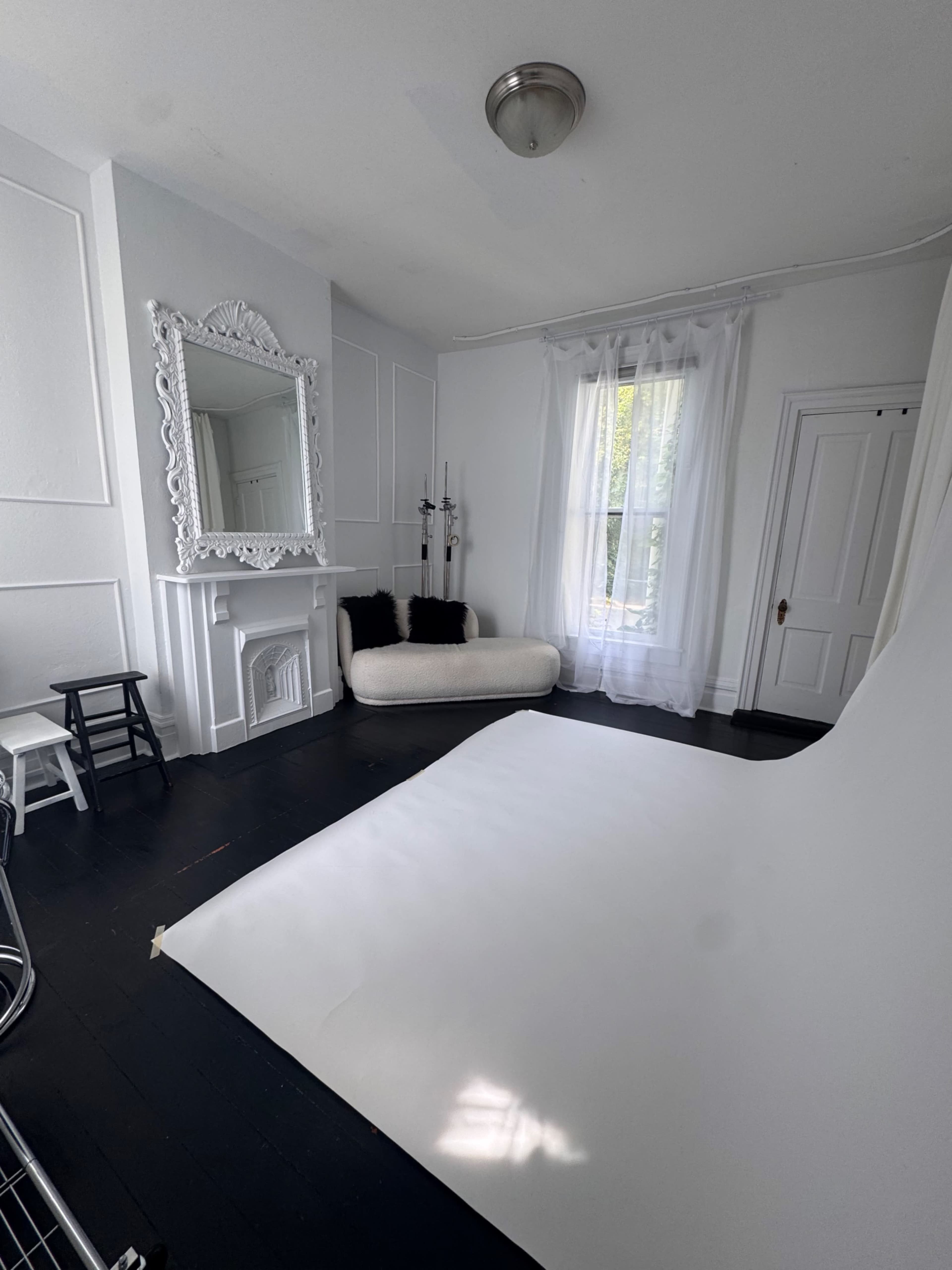 The image shows a bright, minimalist photography studio with a large white backdrop, a vintage mirror, and a small couch near a window.