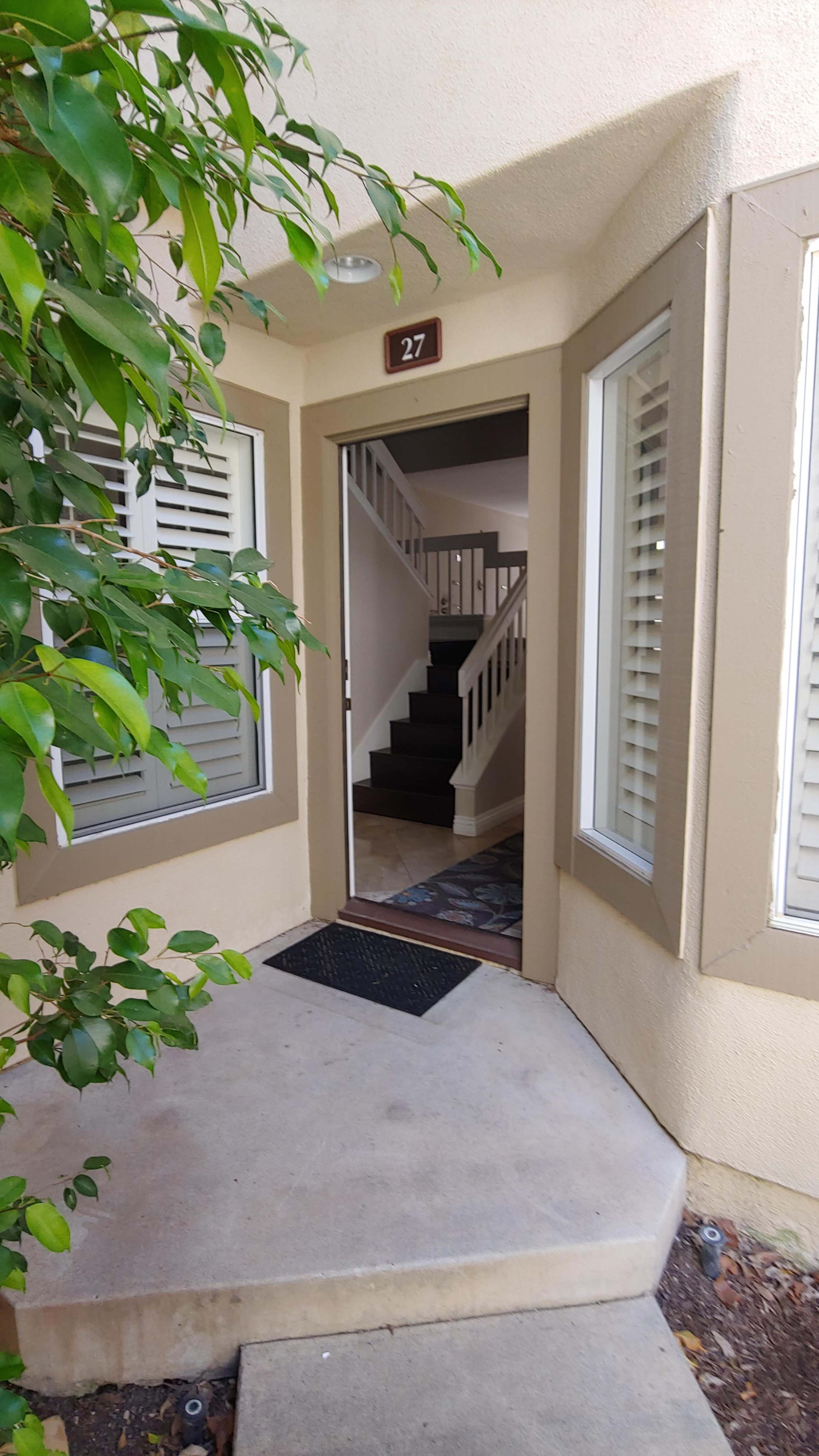 A doorway with a welcome mat leads into a stairwell, framed by potted greenery and adjacent windows.