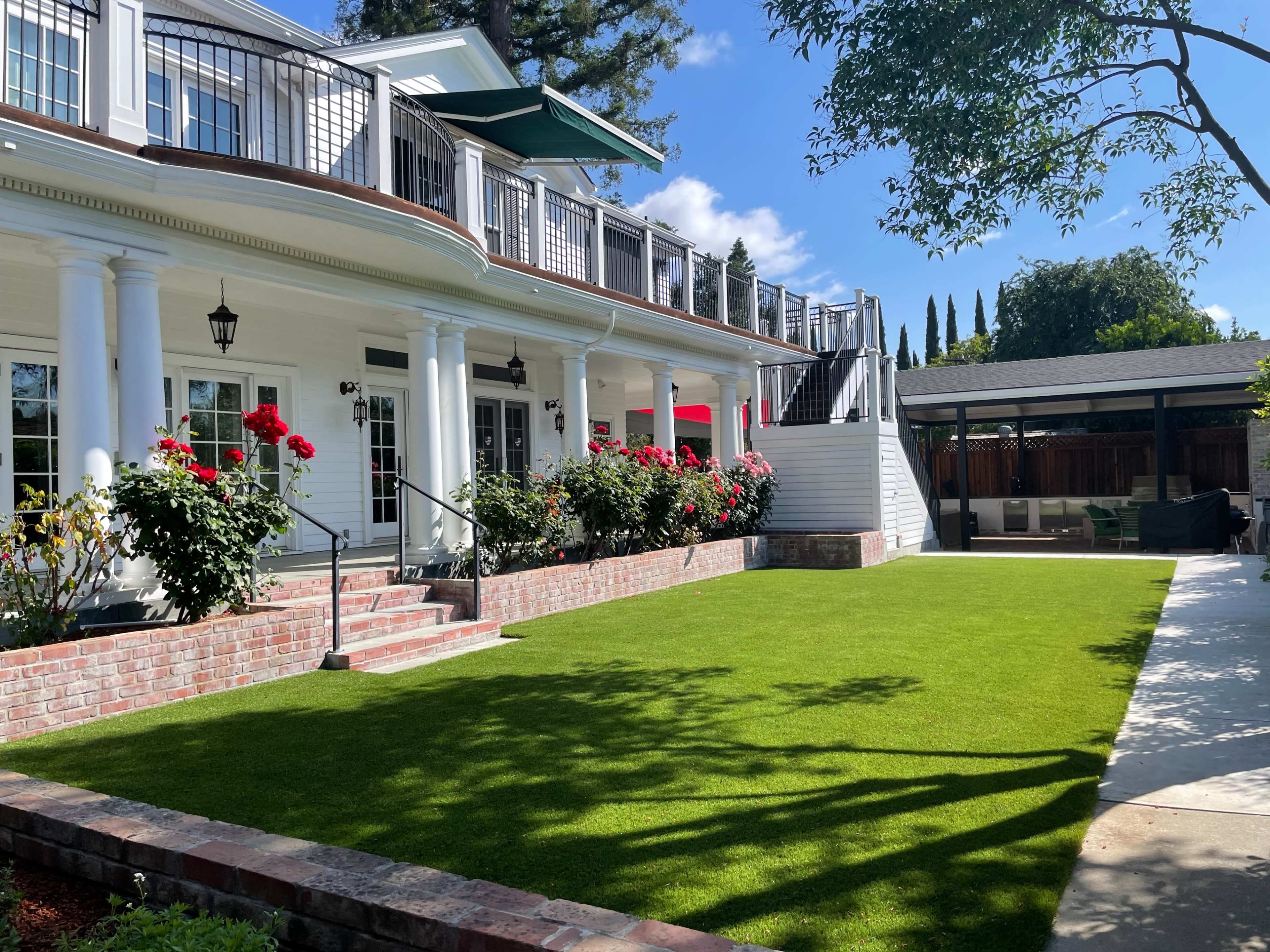 A large white house with a landscaped yard featuring green grass, rose bushes, and a staircase leading to a balcony.