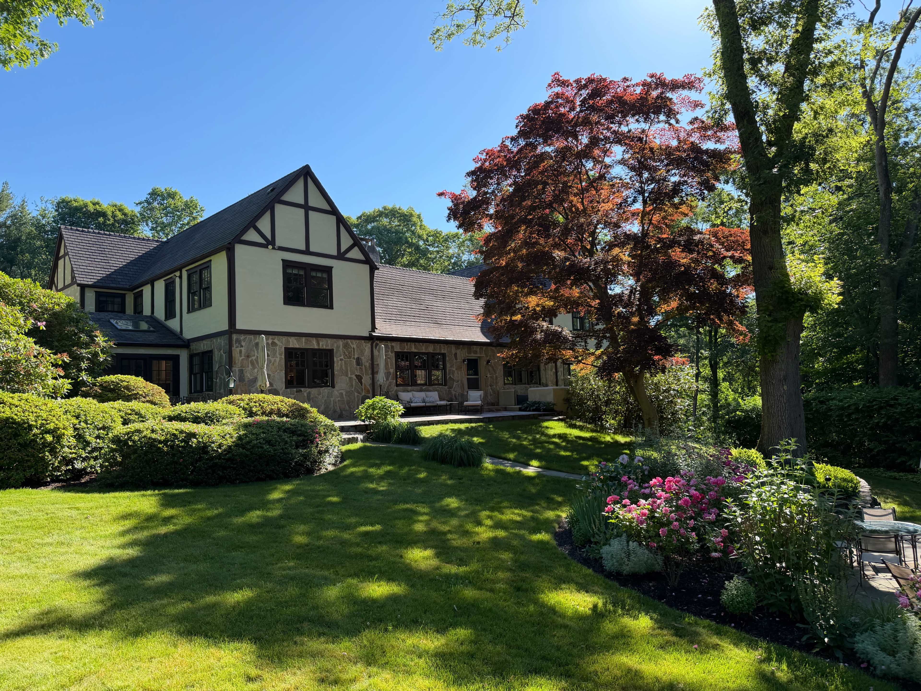 A two-story house with a stone facade and a steeply sloped roof is surrounded by lush greenery and colorful flower beds in a sunny backyard.