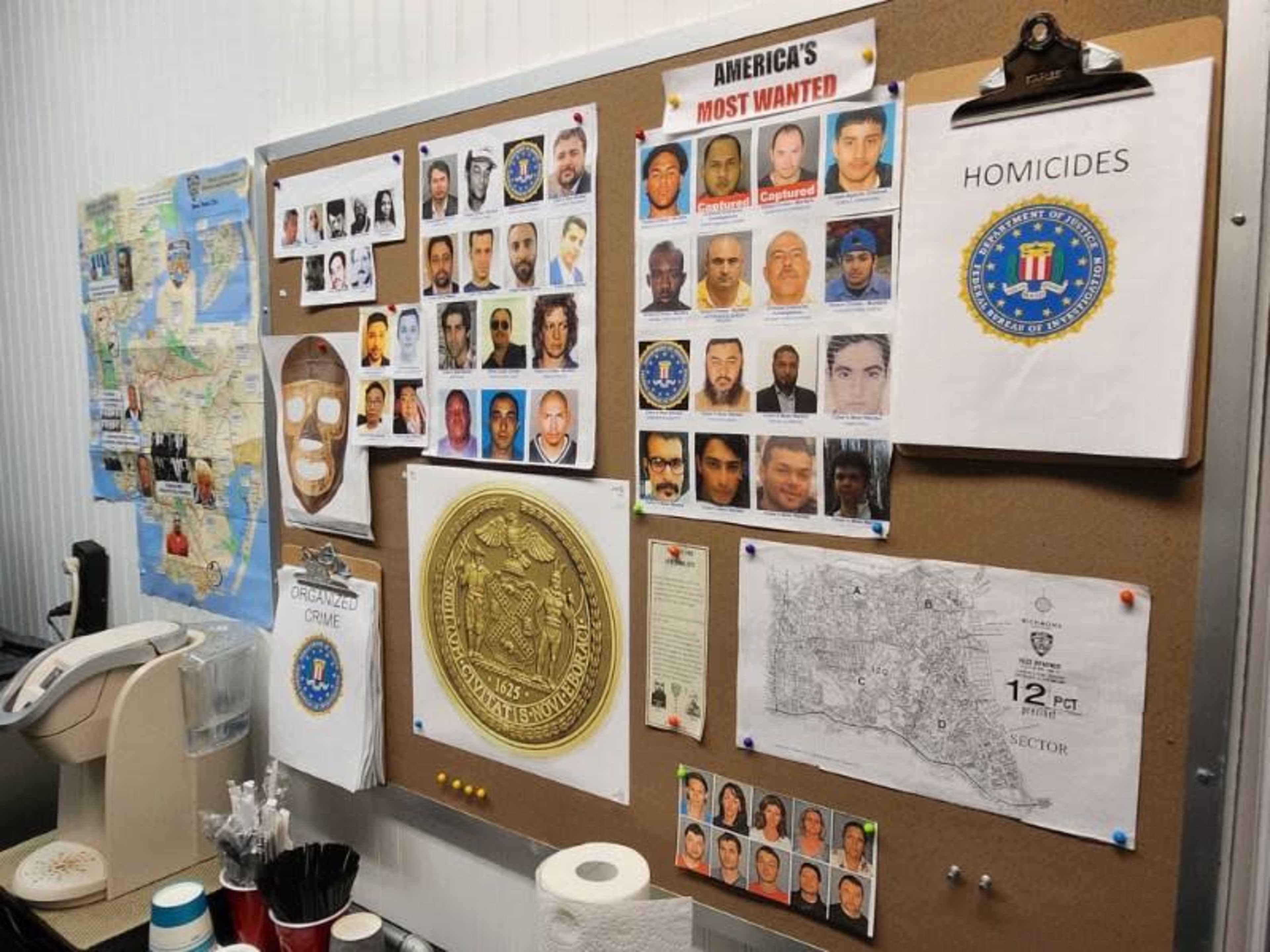A bulletin board displays a collection of wanted posters, maps, and documents related to criminal activity and homicides.