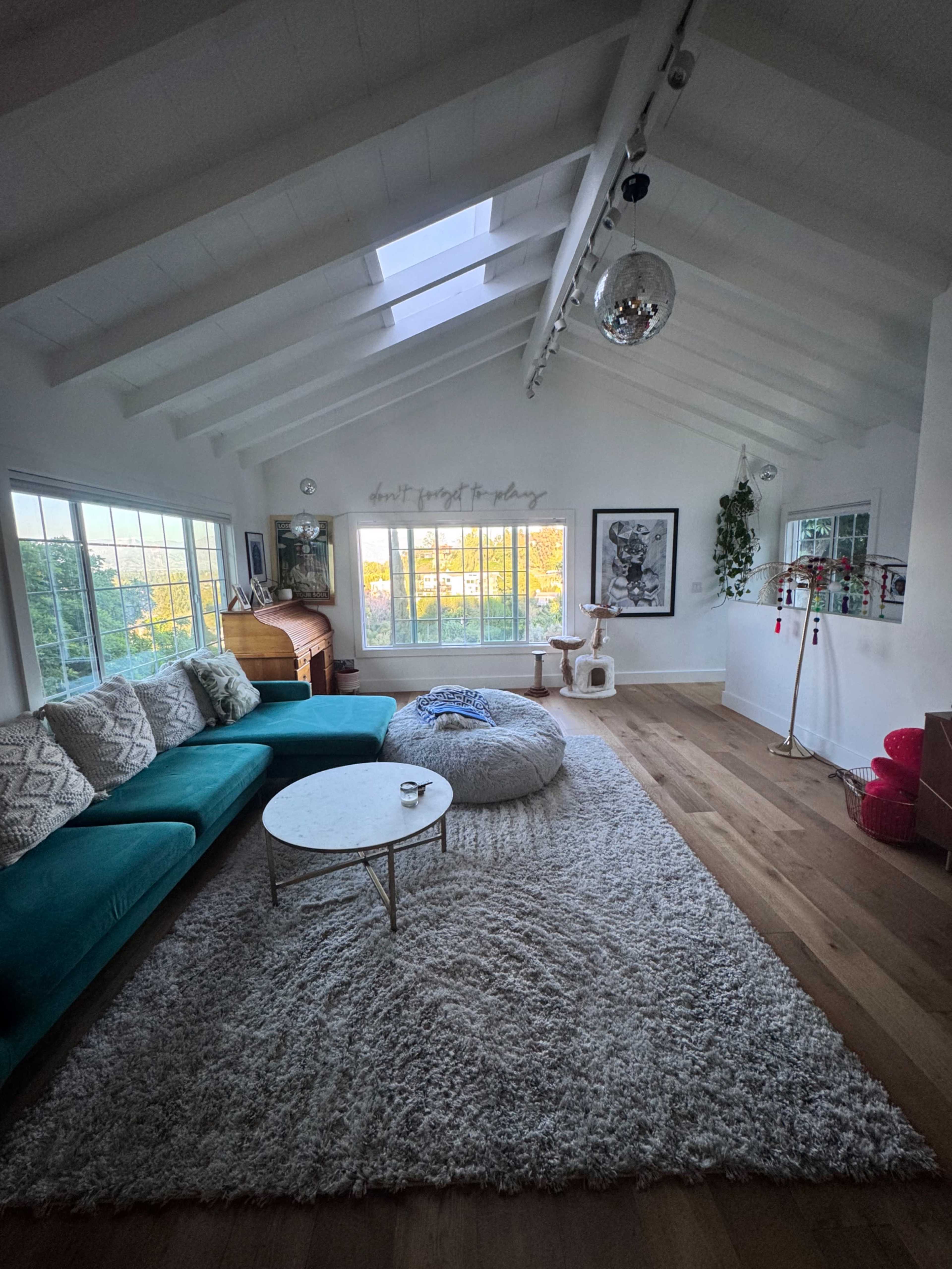 The image shows a bright living room with a sectional couch, a round coffee table, a large area rug, and large windows allowing natural light to fill the space.