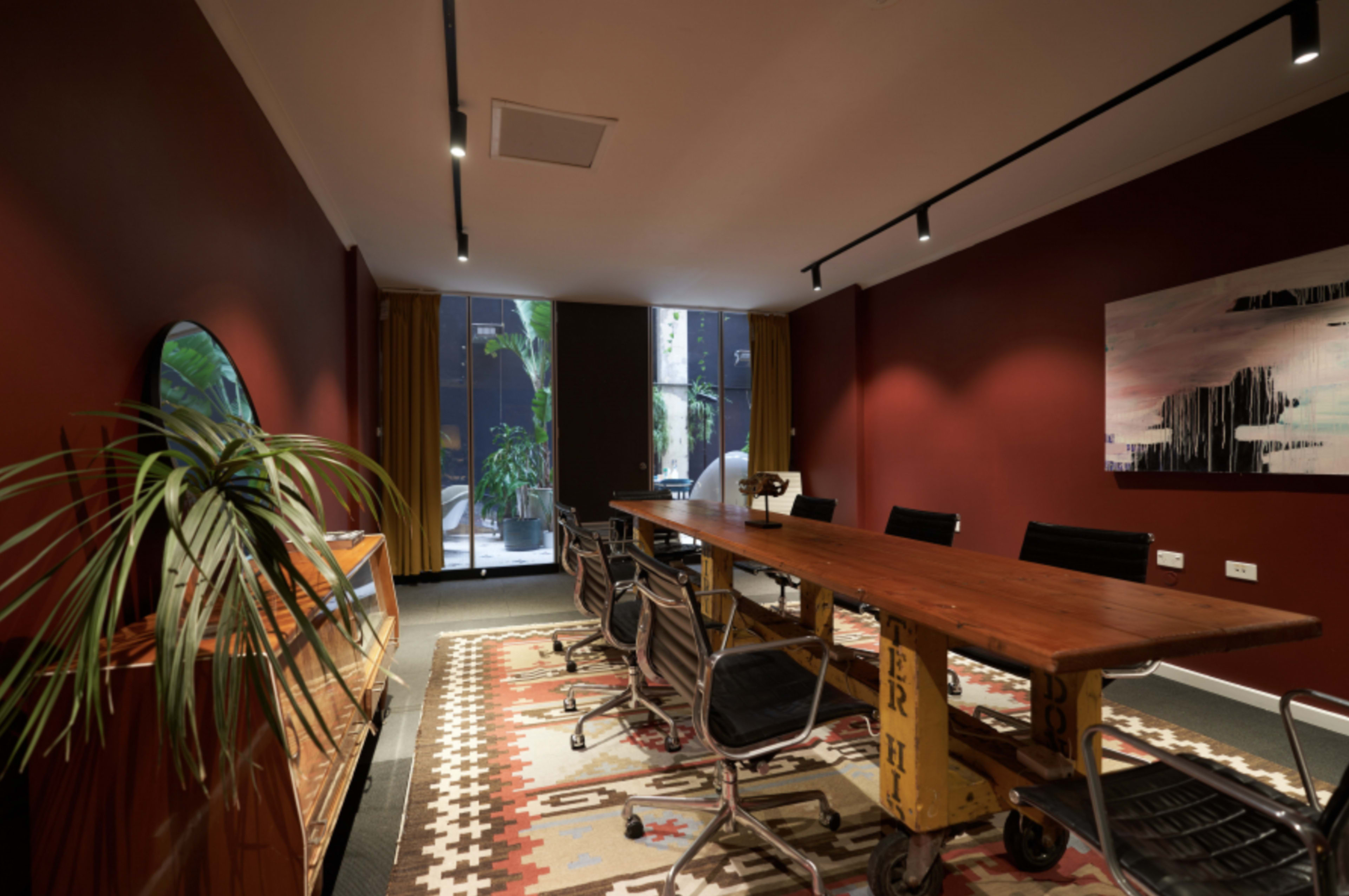 A modern conference room with a long wooden table, black chairs, large windows, and decorative plants along with a patterned rug on the floor.