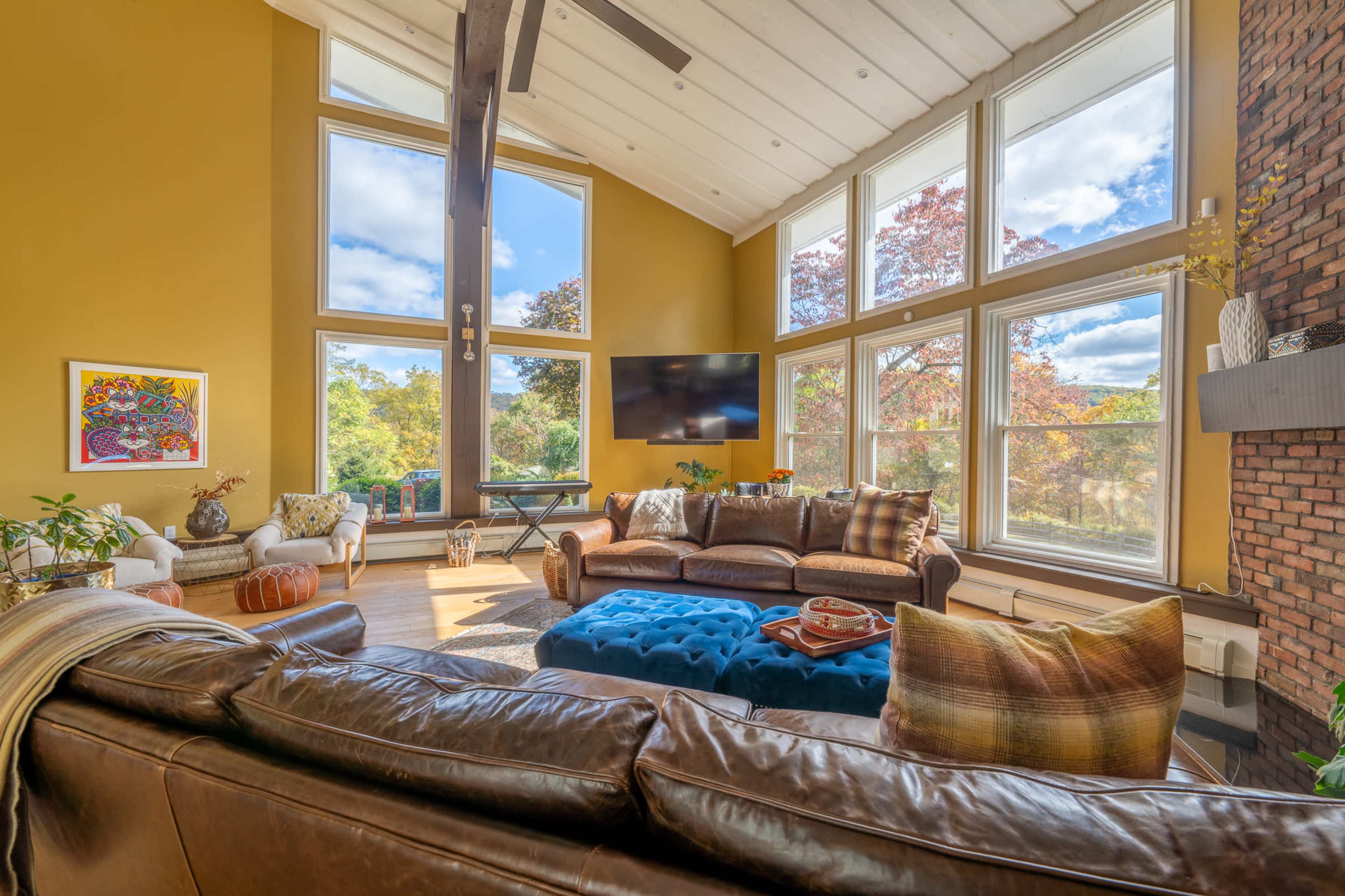 The image shows a spacious living room with large windows, a sectional leather sofa, and a coffee table in the center, featuring a view of greenery outside.