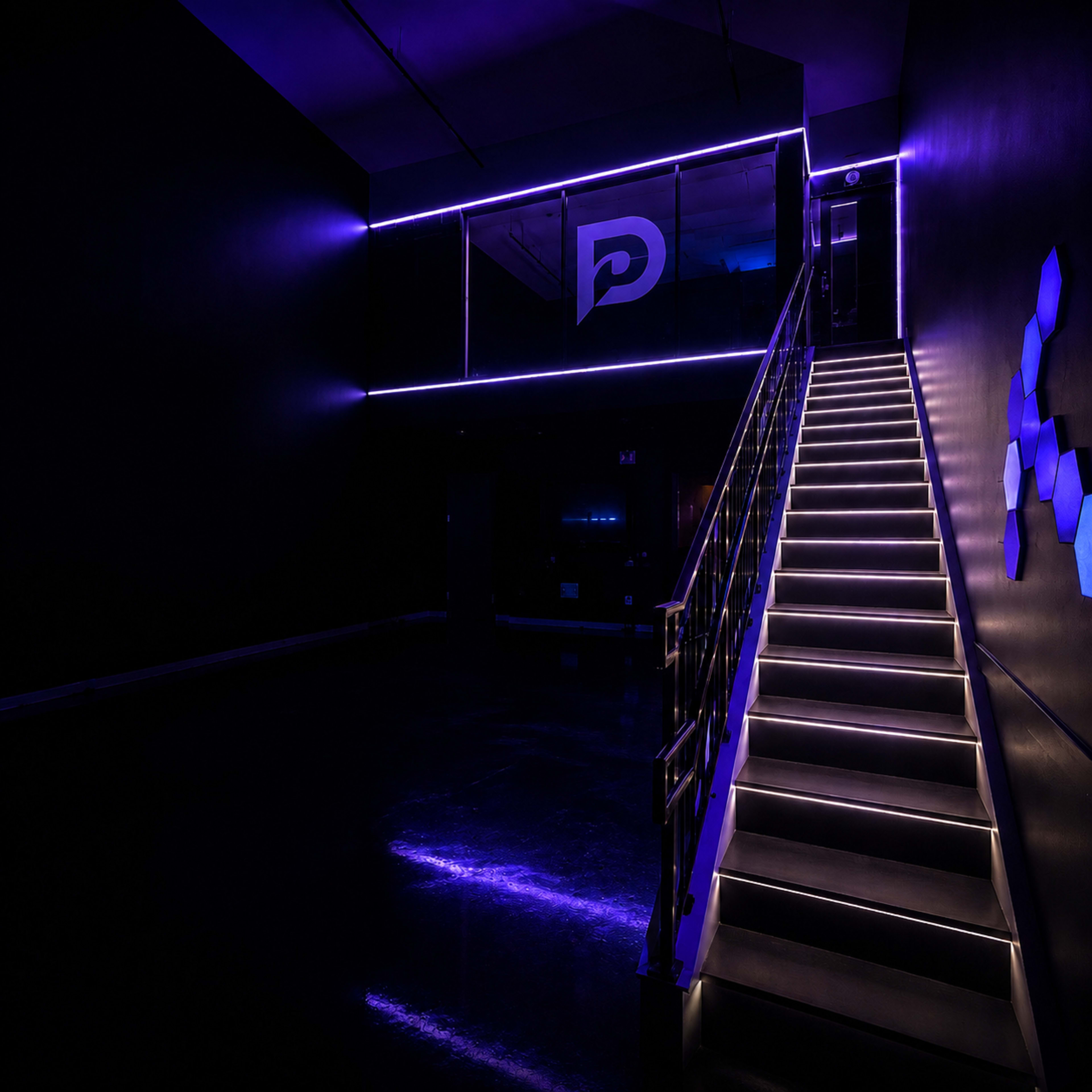 The image shows a dimly lit staircase with illuminated steps leading to a second floor, featuring purple lighting and a large logo on a glass wall.