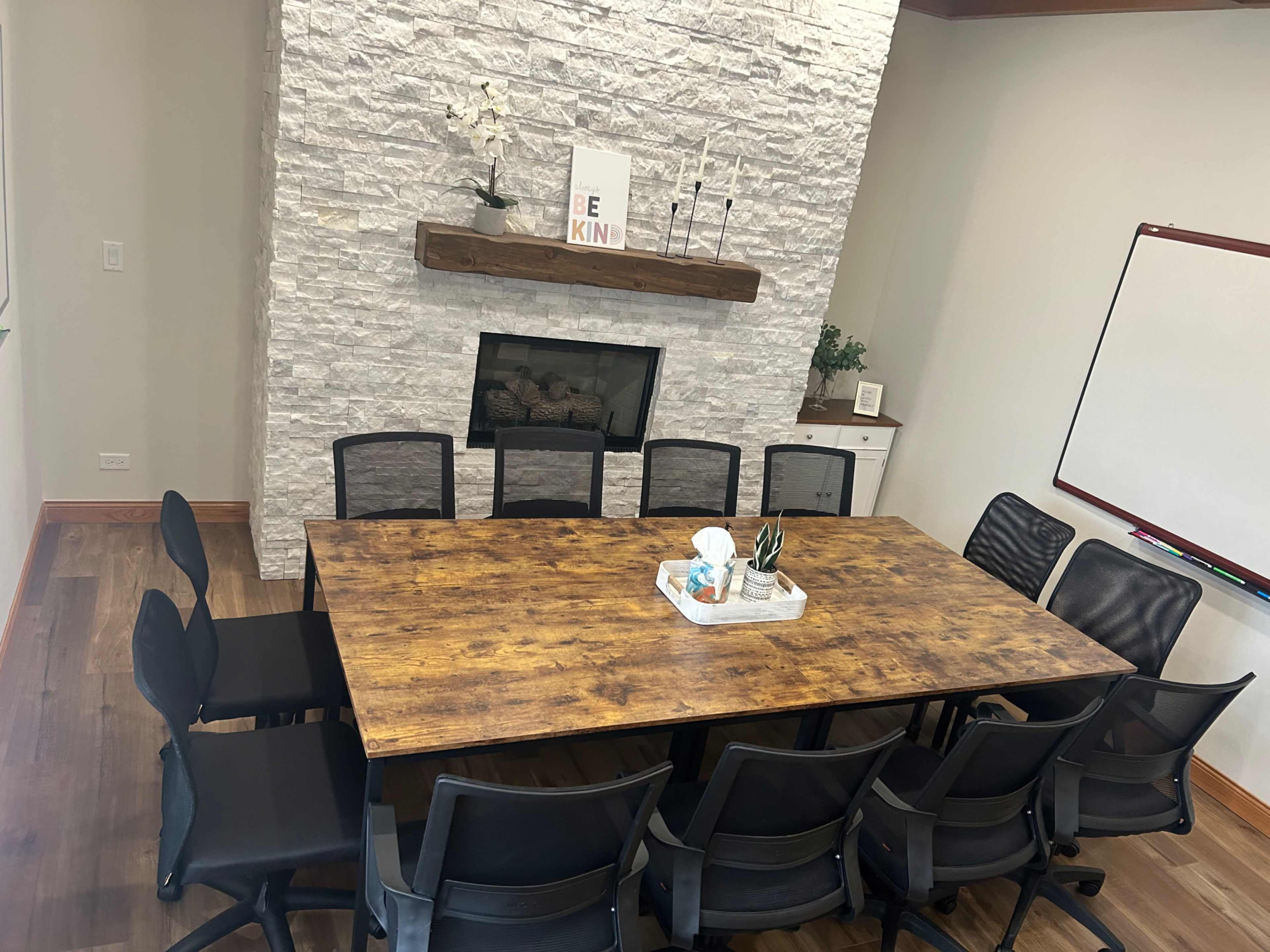 A large wooden table with black chairs surrounds it in a conference room featuring a stone fireplace and a whiteboard.
